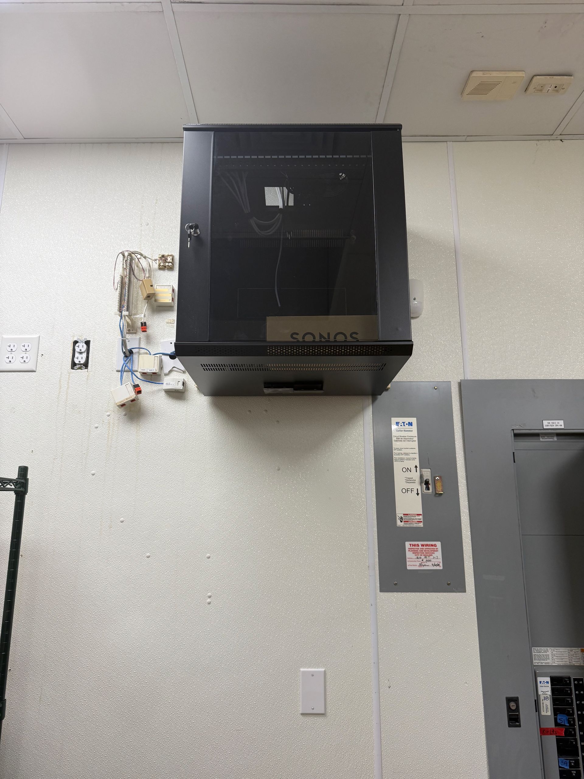 A wall-mounted black server rack with glass door, electrical components, and a gray panel.