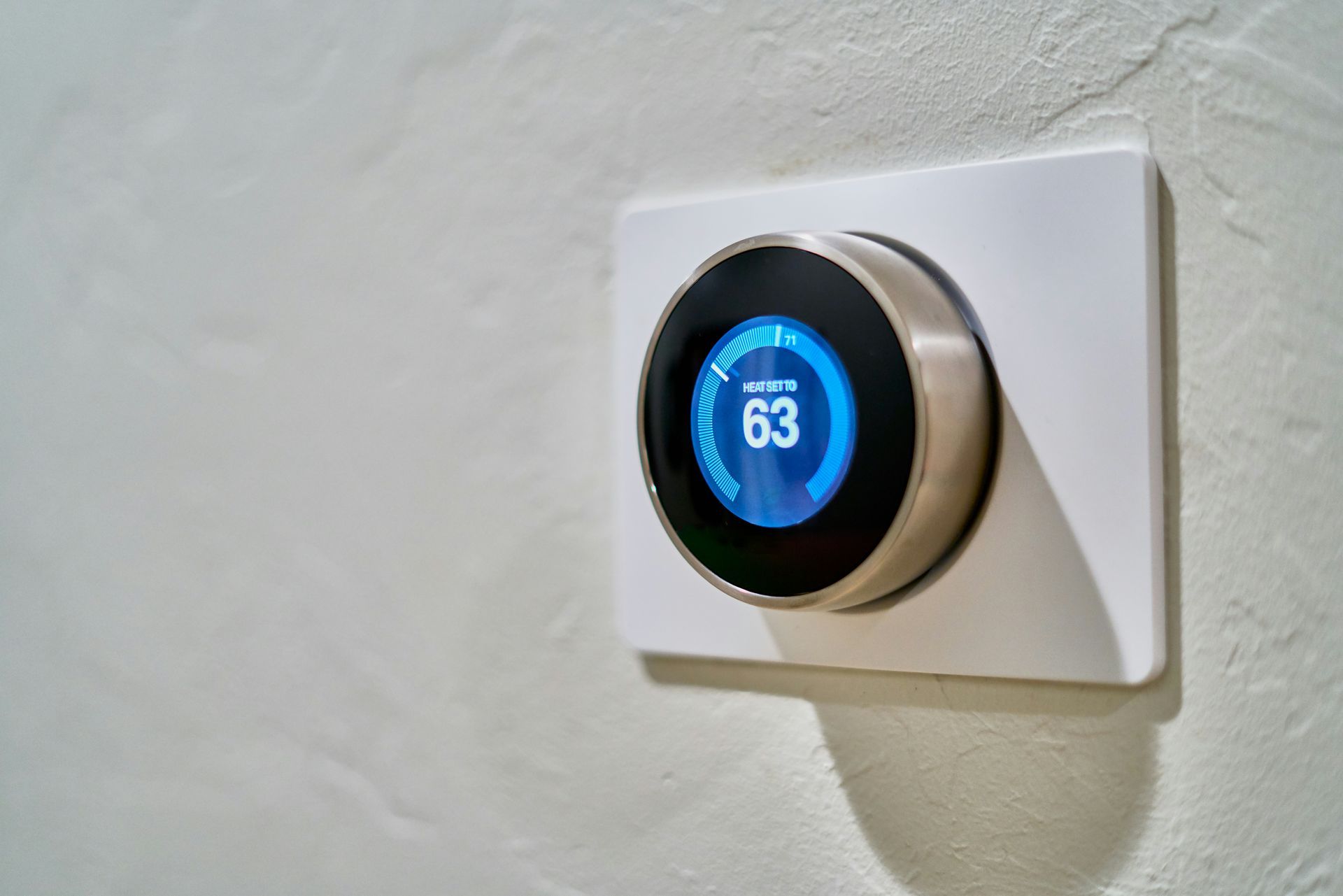 Smart thermostat on a white wall, displaying 63 degrees. Round, metallic design with a blue screen.