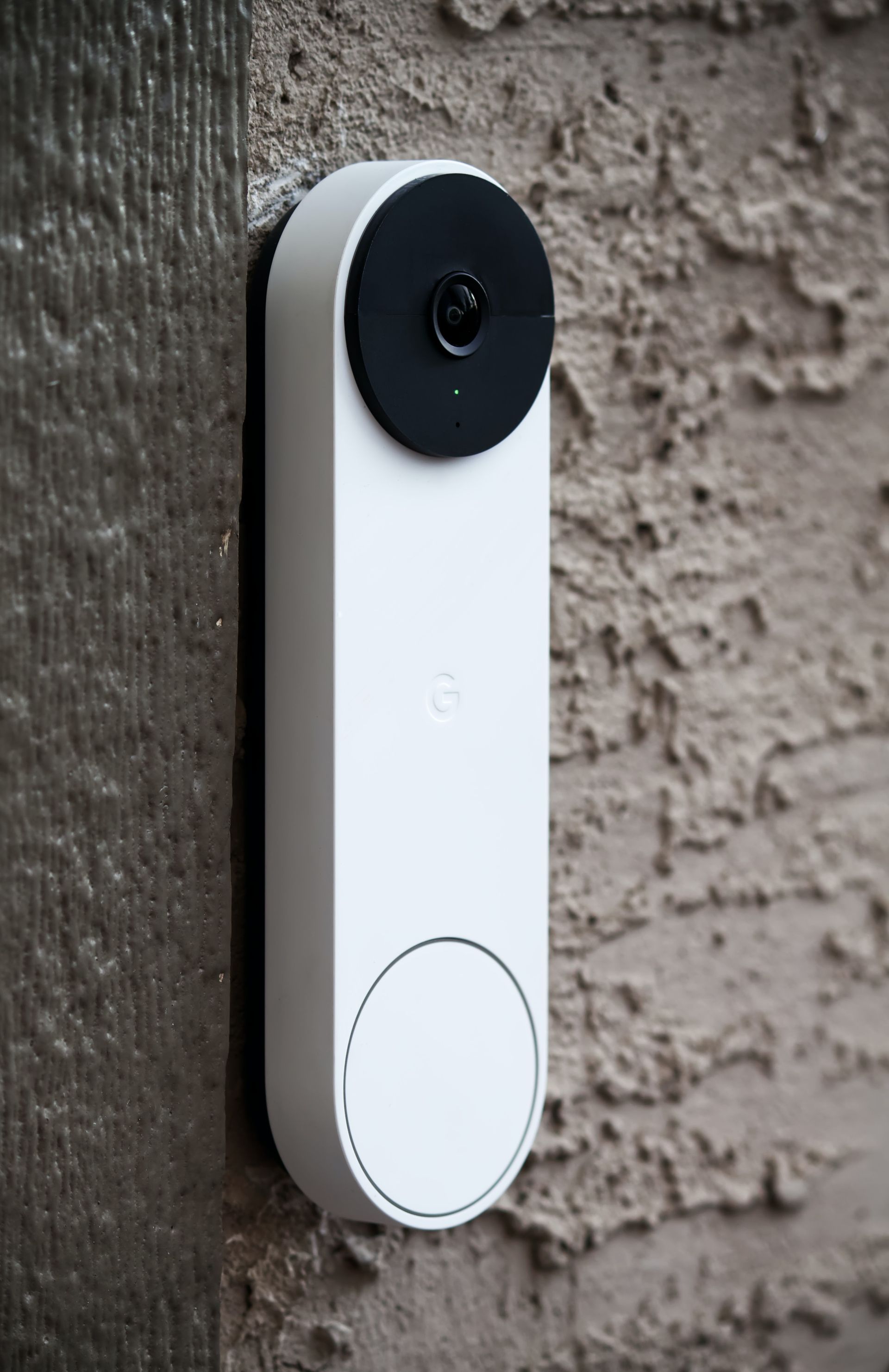 White smart doorbell mounted on a stucco wall, with a black camera lens and a circular button.