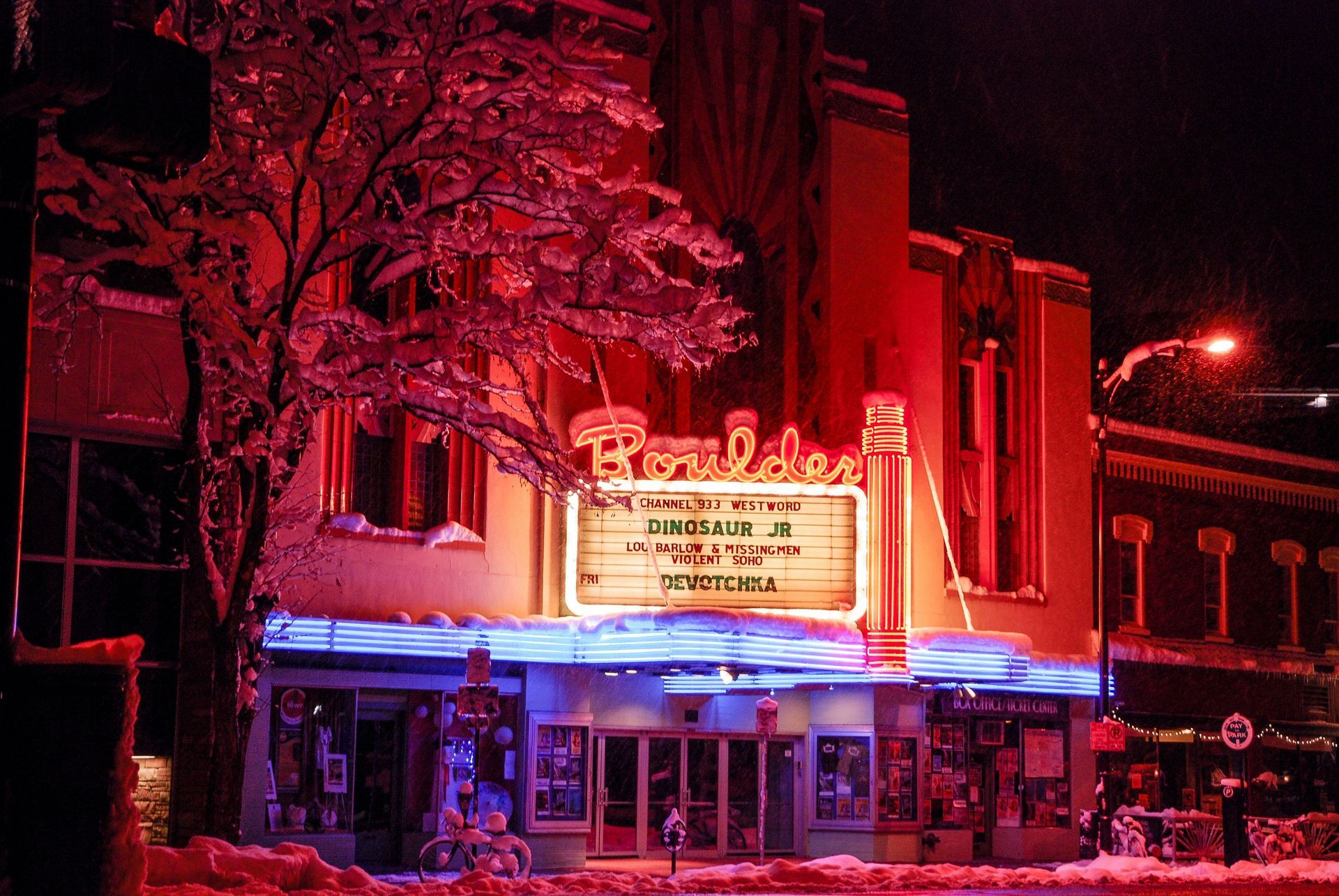 Boulder Theater at night, snow-covered, lit with red and blue neon, marquee displays a show.