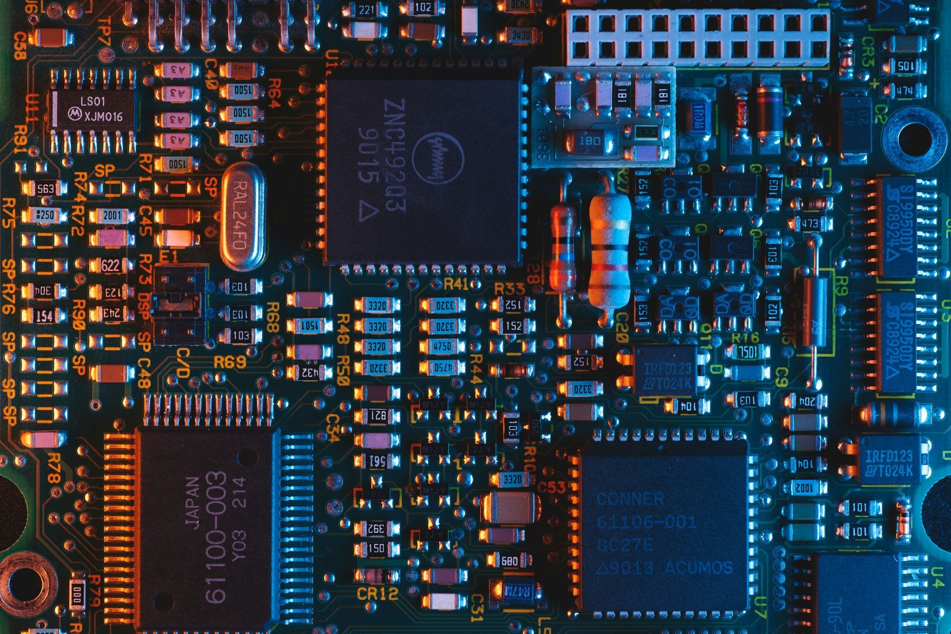 Close-up of a green circuit board with various electronic components; lit with orange and blue light.