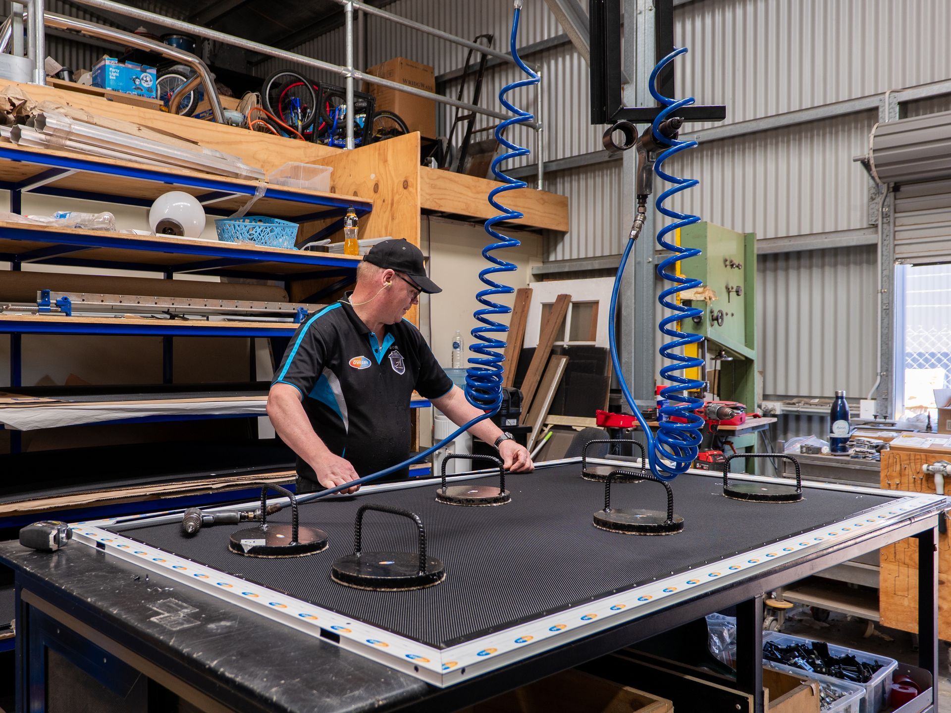 Photograph of Crimsafe Authorised Licensee in a workshop with a large flat surface featuring four suction cups attached to a black material Photograph of Crimsafe Authorised Licensee in a workshop with a large flat surface featuring four suction cups attached to a black material