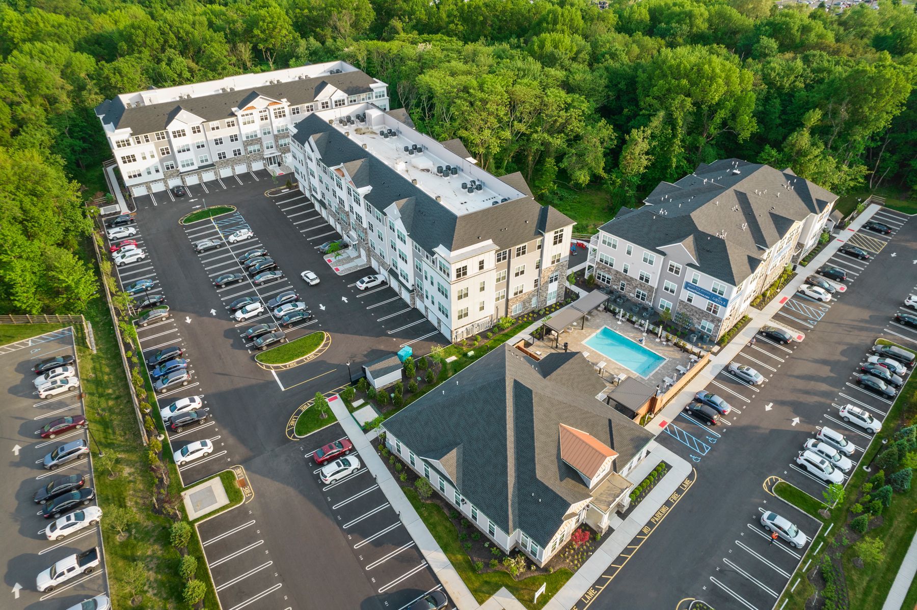 Aerial view of an apartment complex with multiple buildings, parking lots, and a pool surrounded by trees.