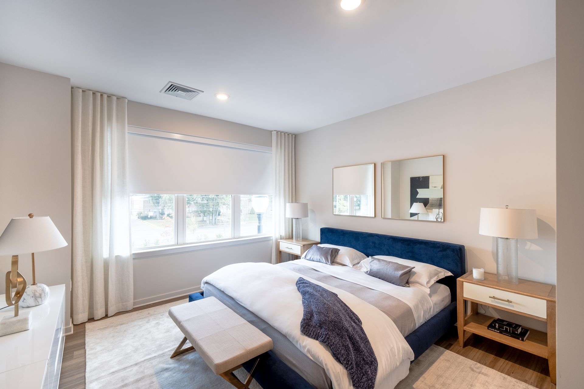 Bedroom with blue bed, cream curtains, bedside tables, and a bench.