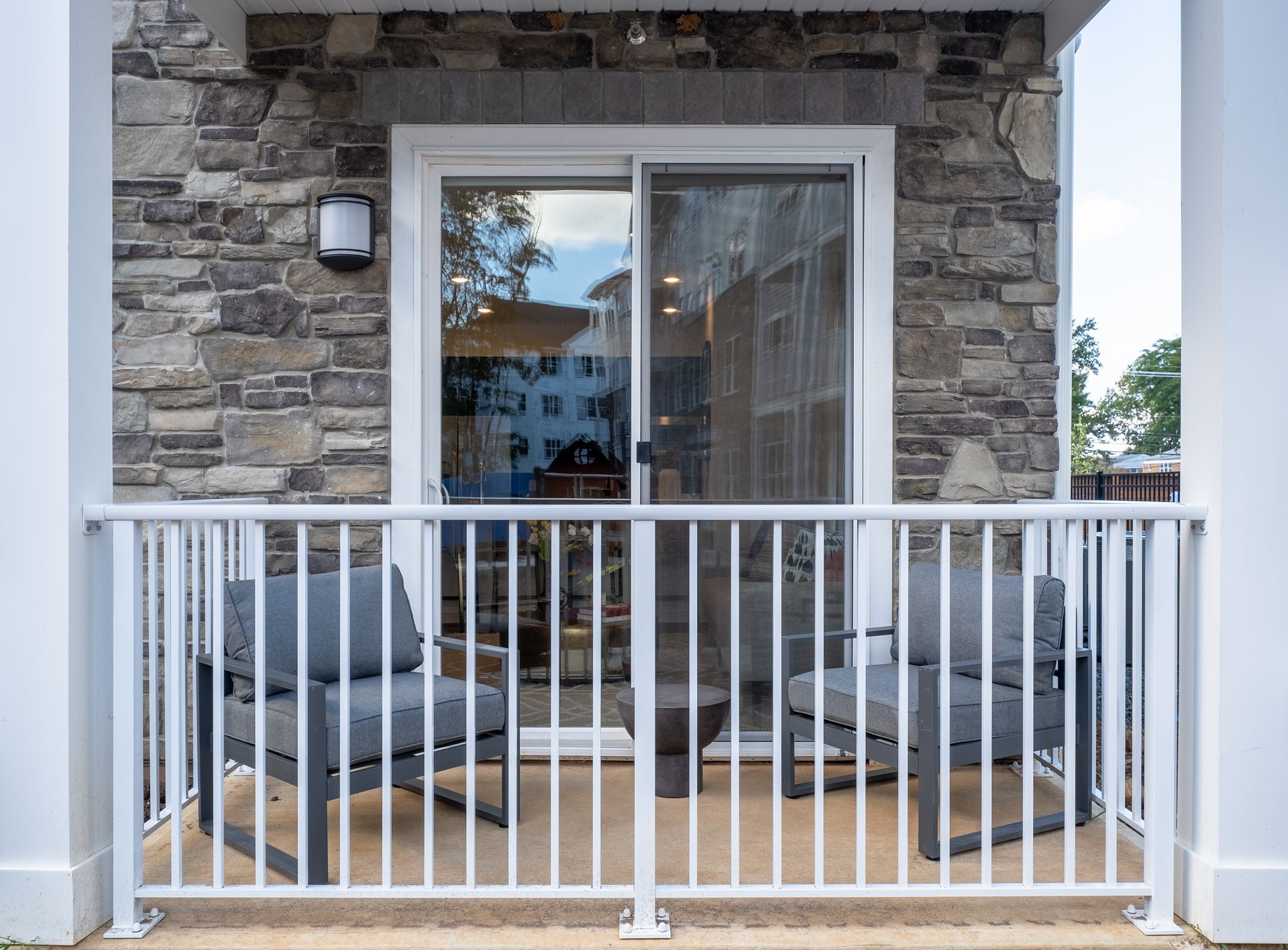 Balcony with two gray chairs and small table, white railing, sliding glass door, stone wall.