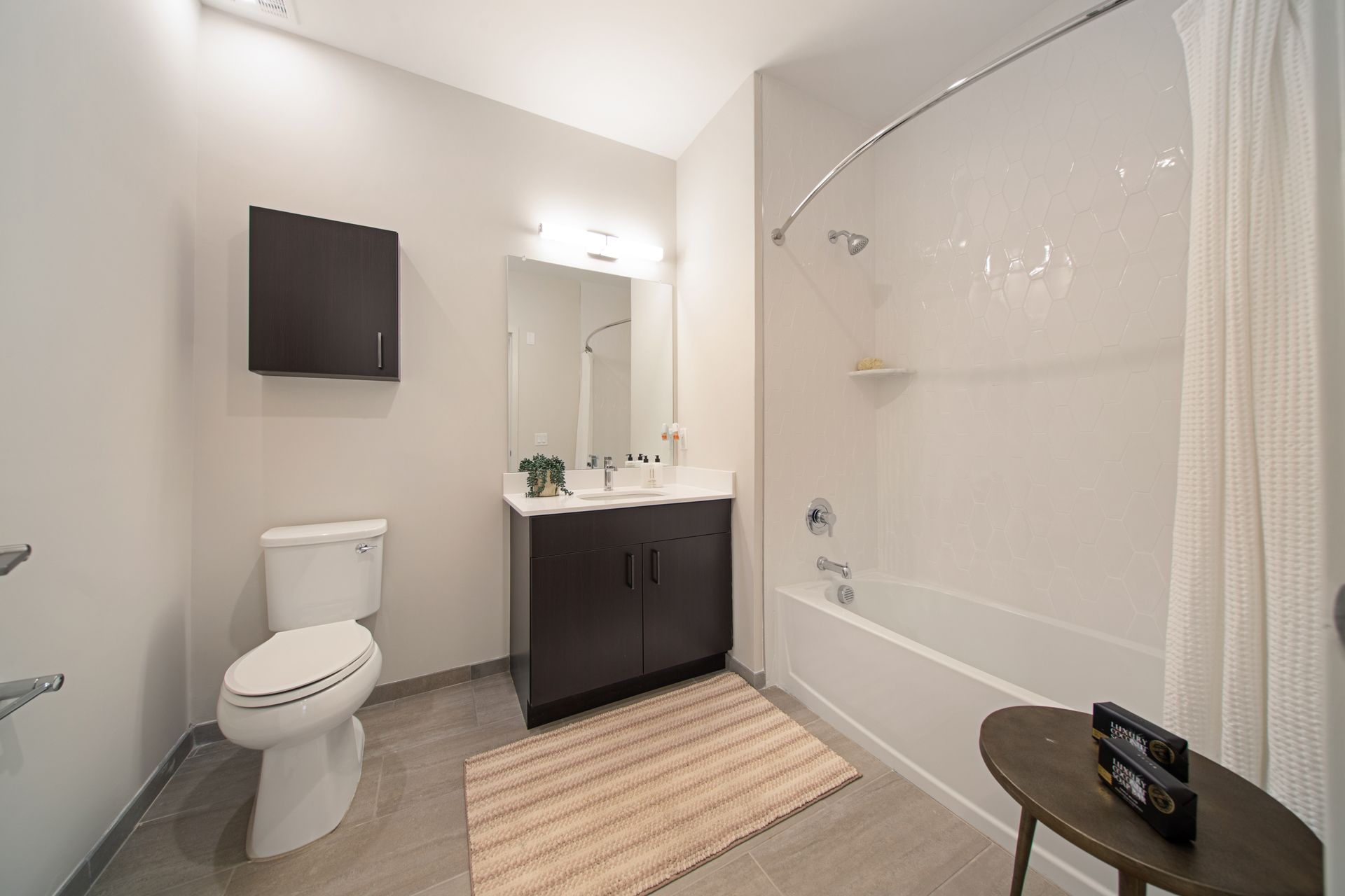 Modern bathroom with white tub, vanity, toilet, and gray flooring. A small rug sits on the floor.