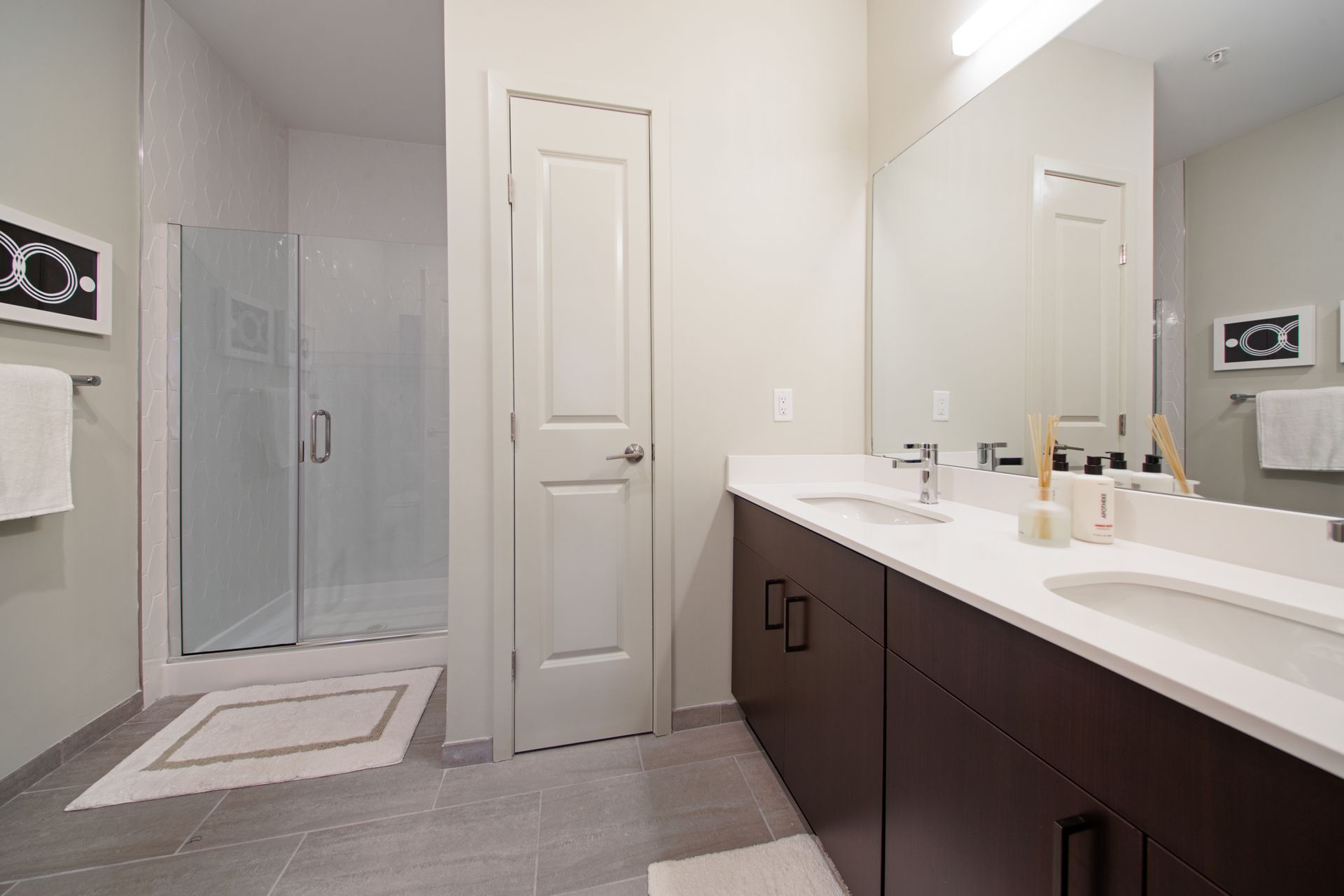 Bathroom with a glass shower, double sink vanity, white door, and gray tile floor.
