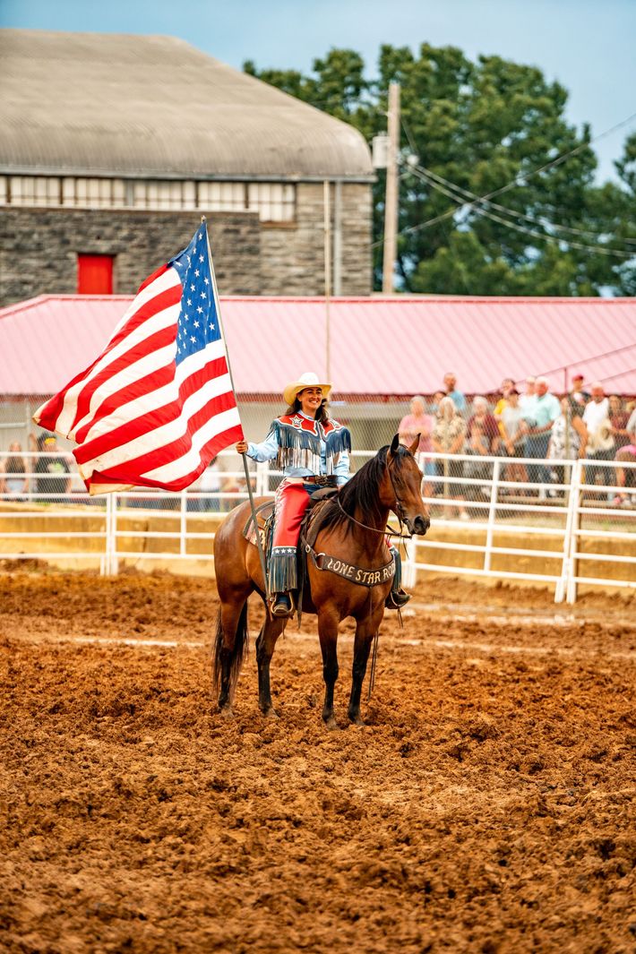 A man is riding a horse in a dirt field while holding an american flag.