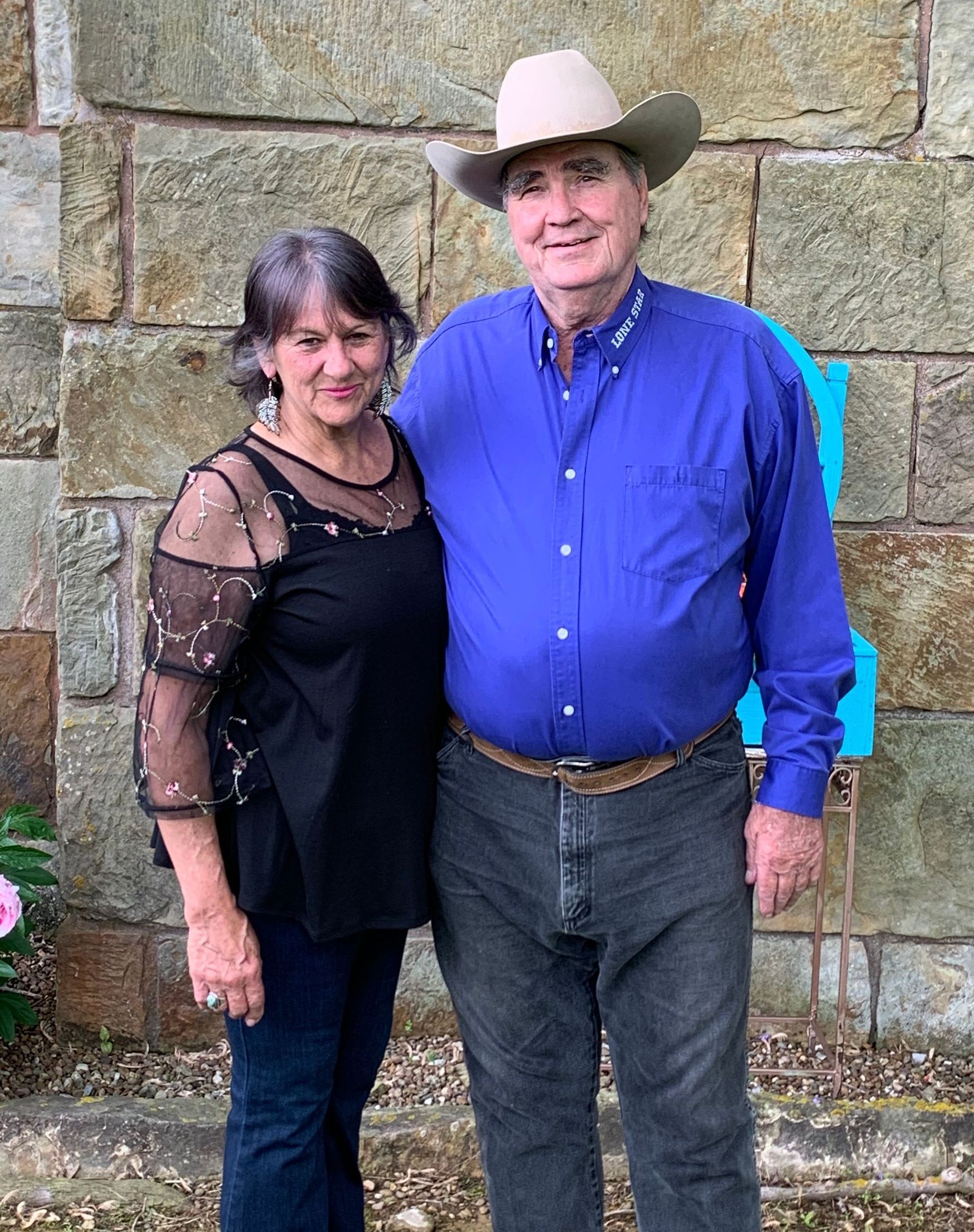 A man and woman are posing for a picture in front of a stone wall . the man is wearing a cowboy hat.