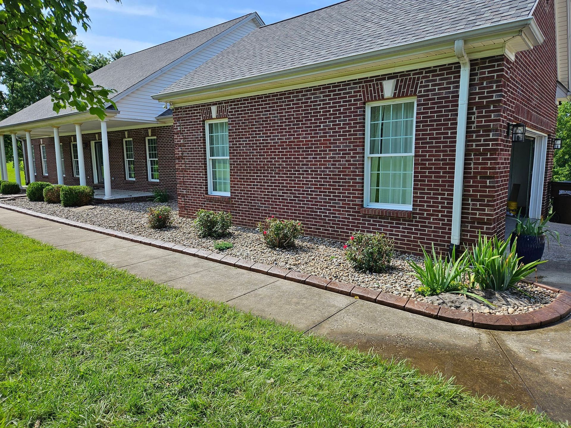 Brick house with a porch, landscaping, and a sidewalk on a sunny day.