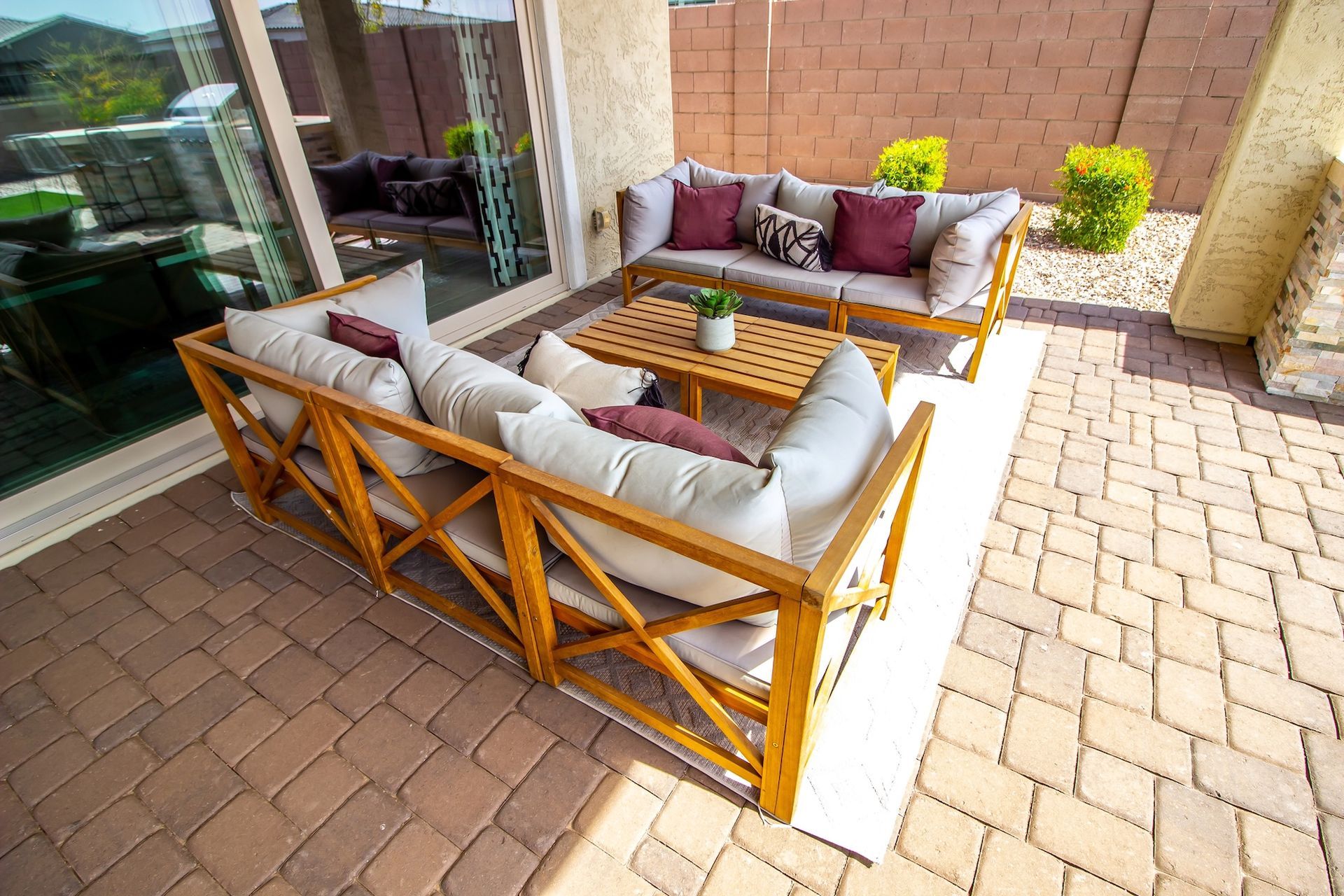 Outdoor patio furniture set on a brick patio. Wooden frames, beige cushions, and burgundy pillows.