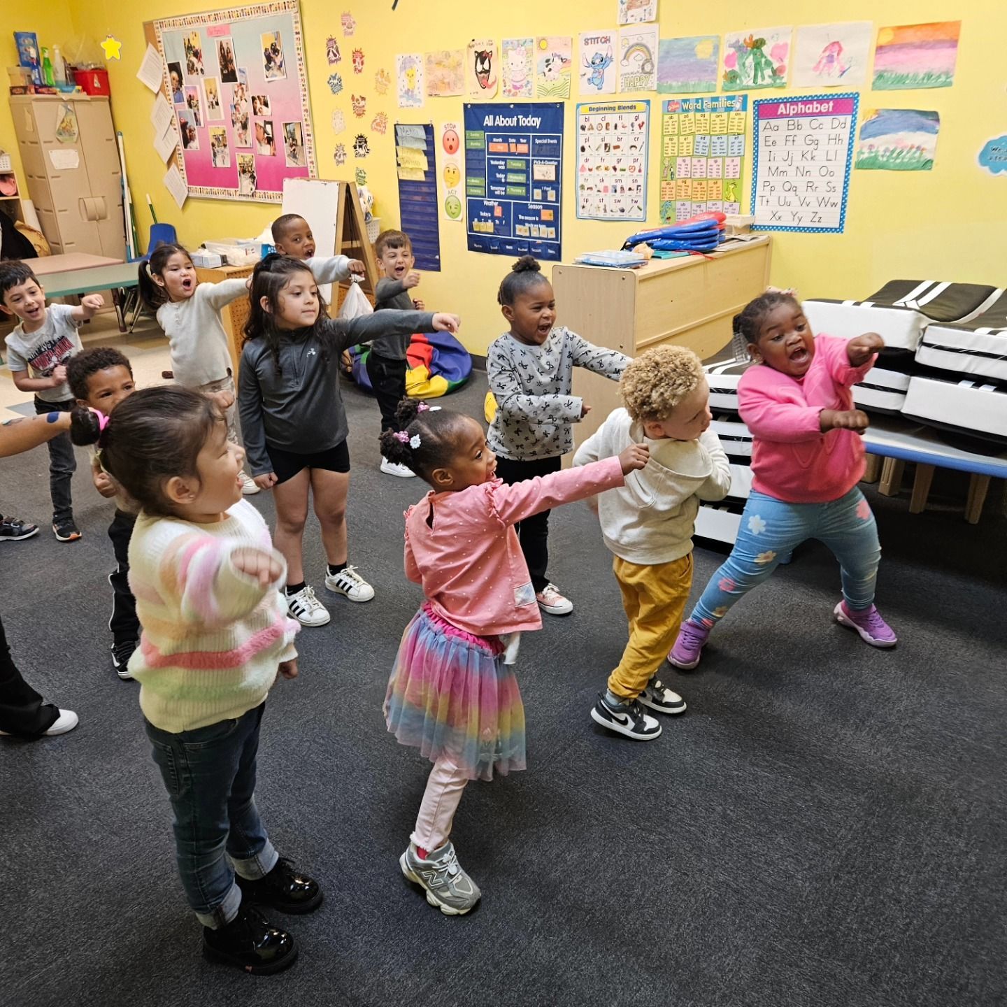 A group of children in a colorful classroom stand in rows, stretching their arms forward while participating in an activity.