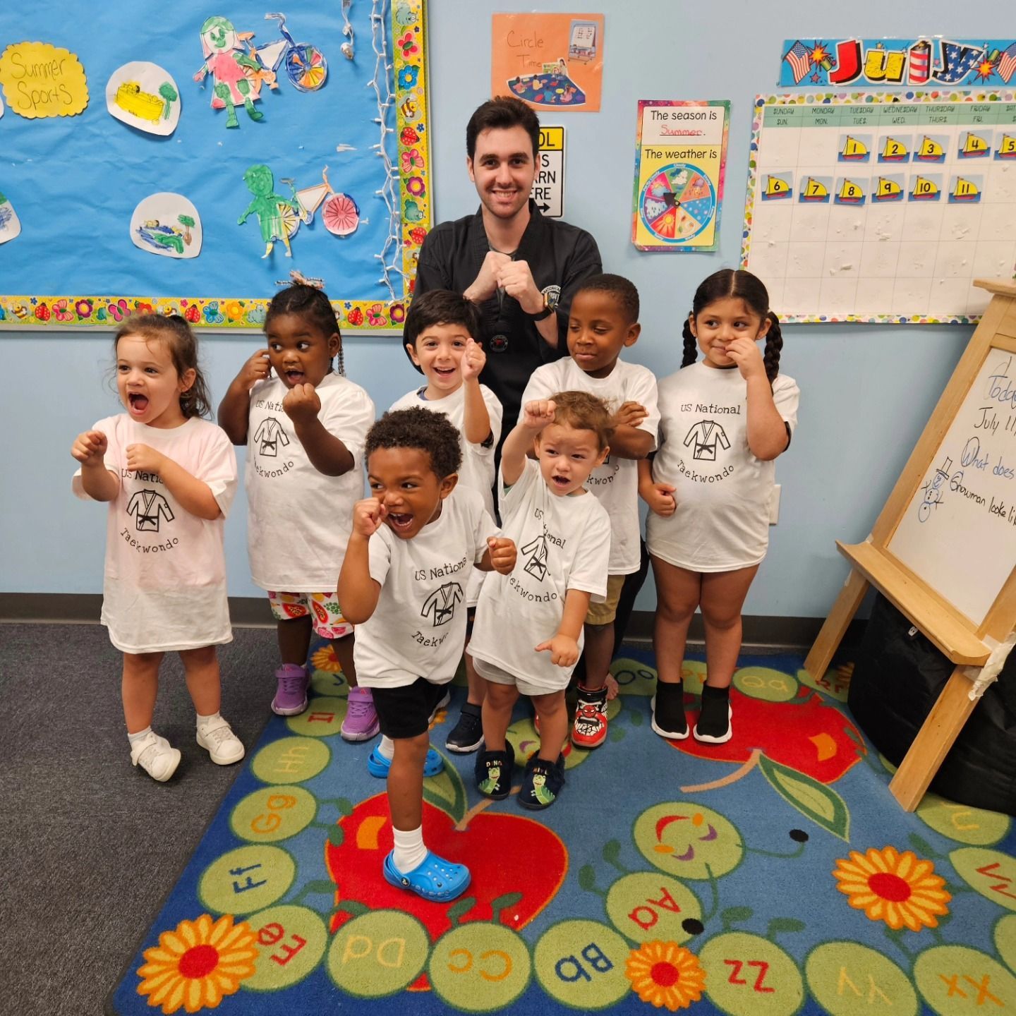 A group of children and a teacher in a classroom making fun faces and gestures in front of a colorful learning rug.
