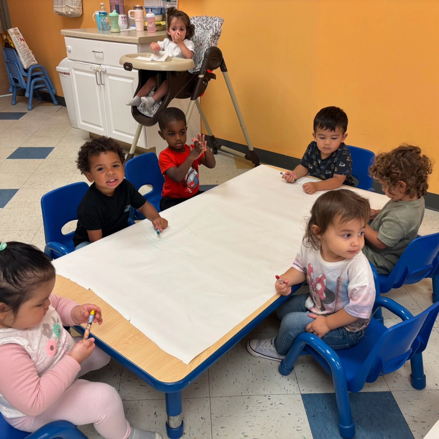 Children sit at a table covered with white paper, with one child in a high chair, in a classroom setting.
