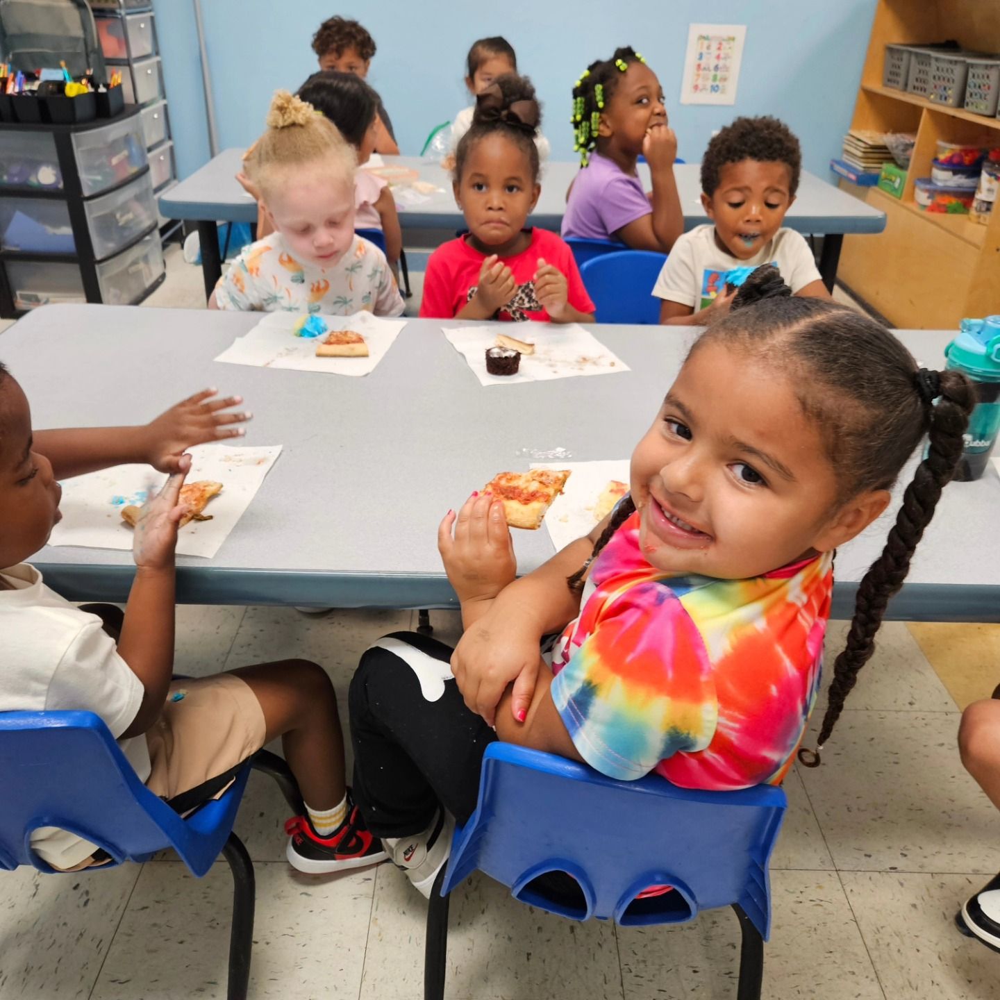 Children sit at tables in a classroom eating pizza, with one smiling toward the camera in a tie-dye shirt.