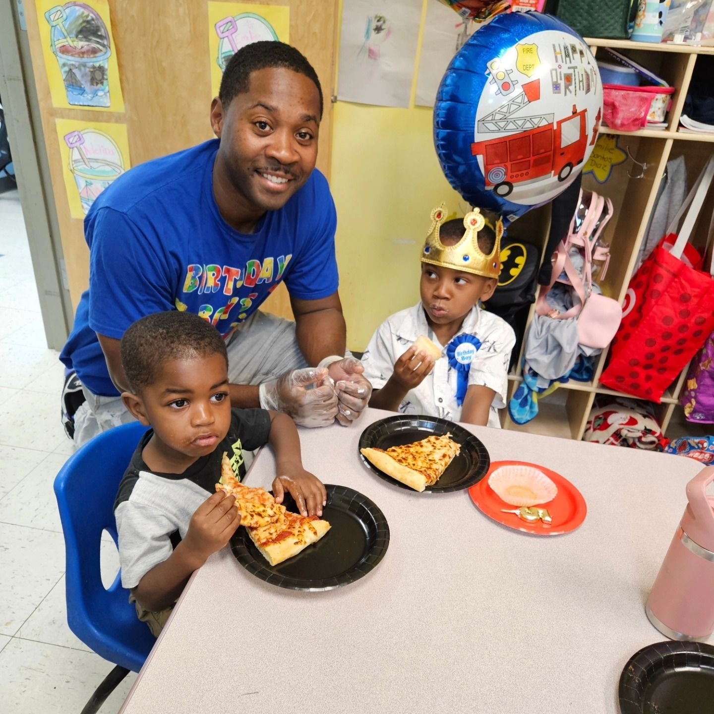 An adult and two children eating pizza at a table in a classroom, with a birthday balloon and party crown present.