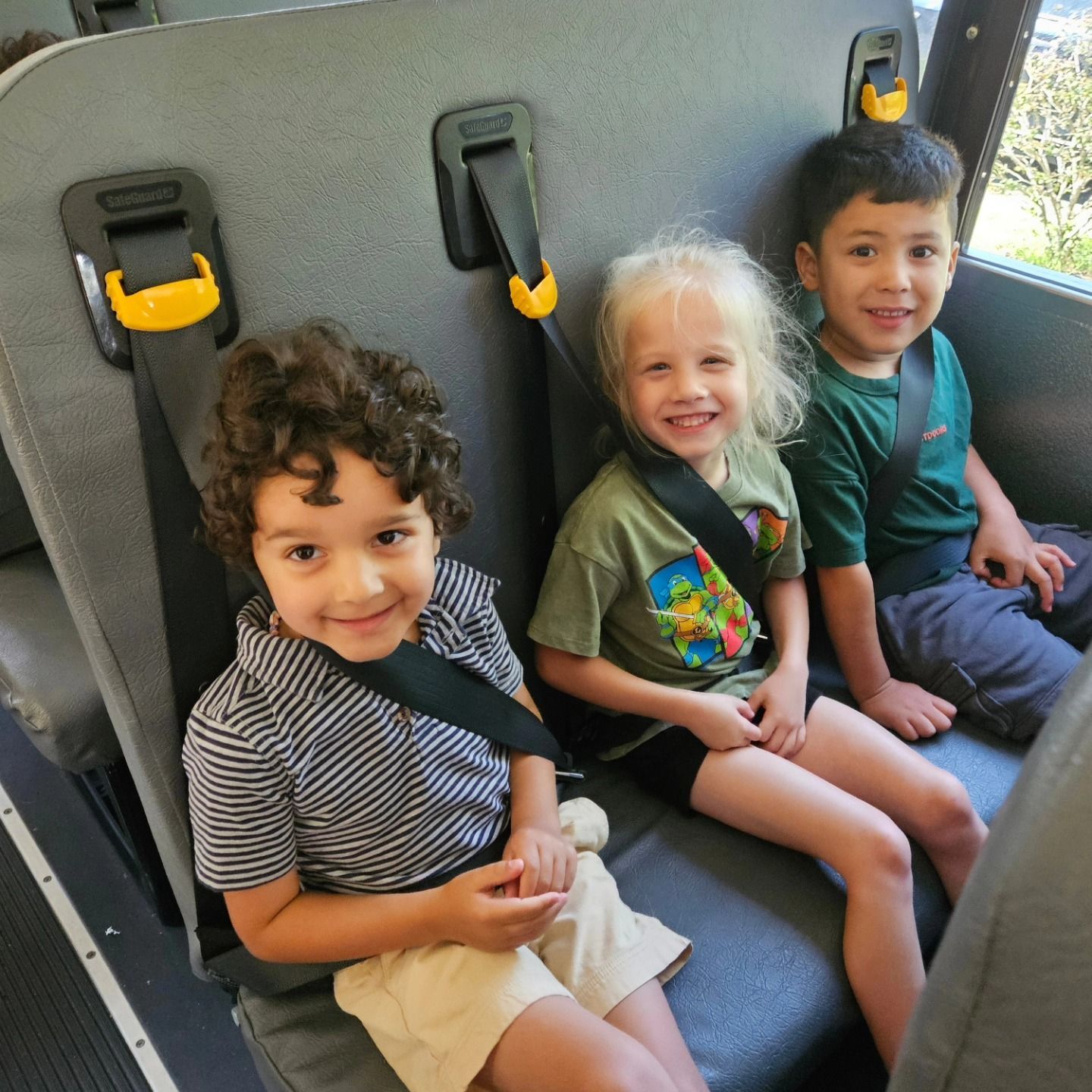 Three children sit side-by-side in a school bus seat, smiling while wearing their seatbelts.
