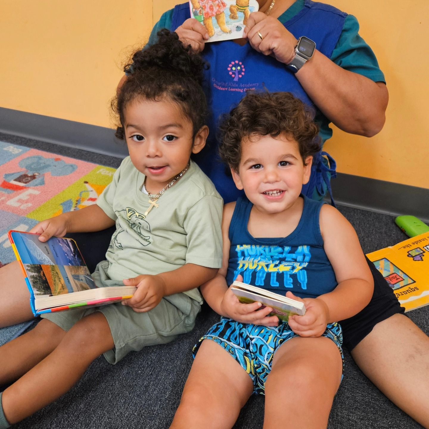 An adult holds a book while two children sit on the floor, each holding a book in a brightly lit indoor space.