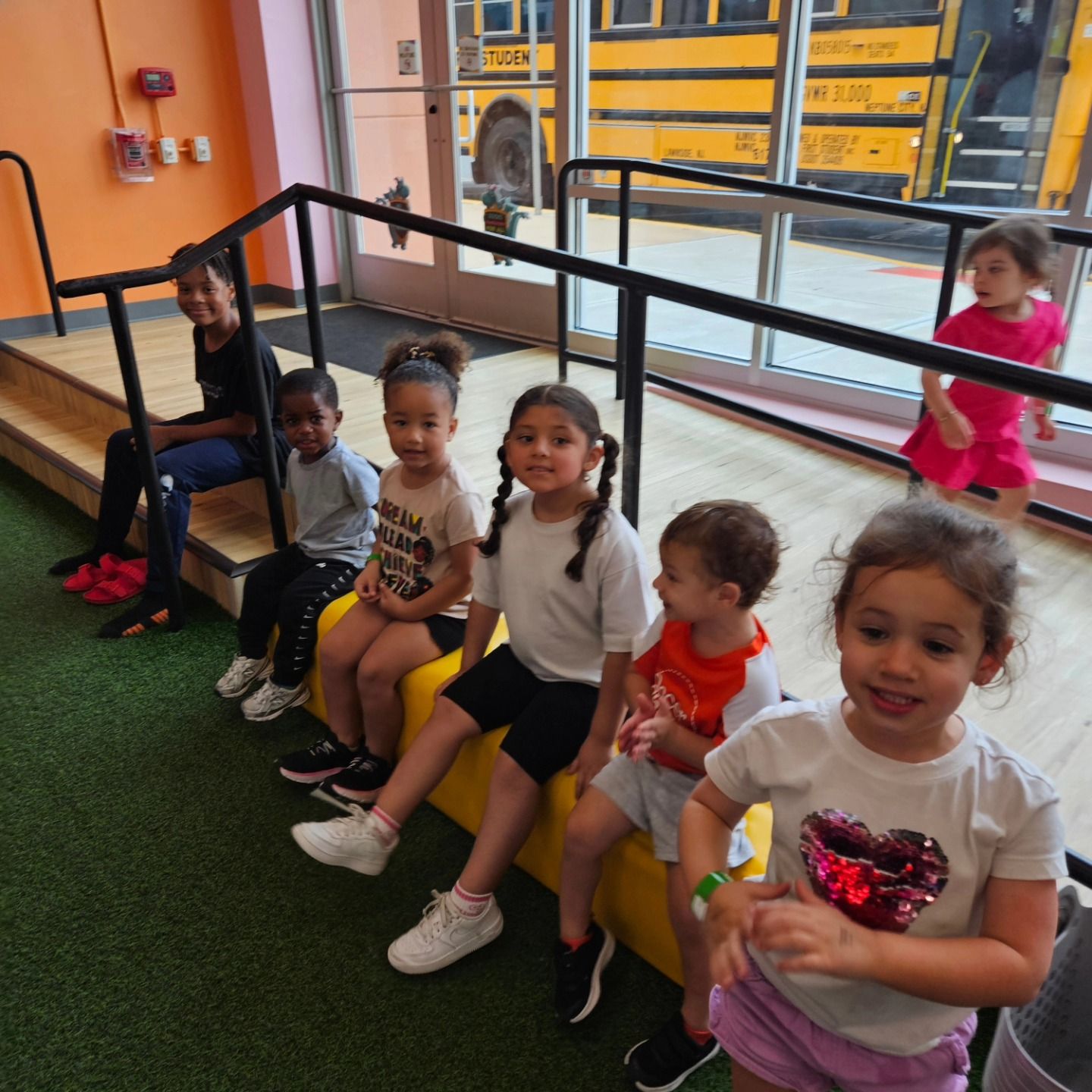 A group of children sit on a yellow bench inside, while one child stands near a window with a school bus outside.