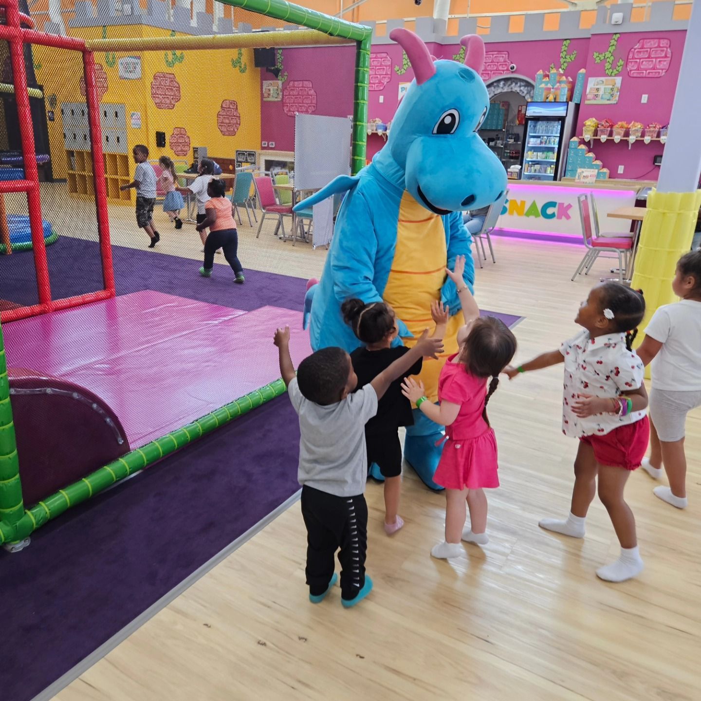 Children interact with a blue dragon mascot in a brightly colored indoor playground with play structures and a snack bar.