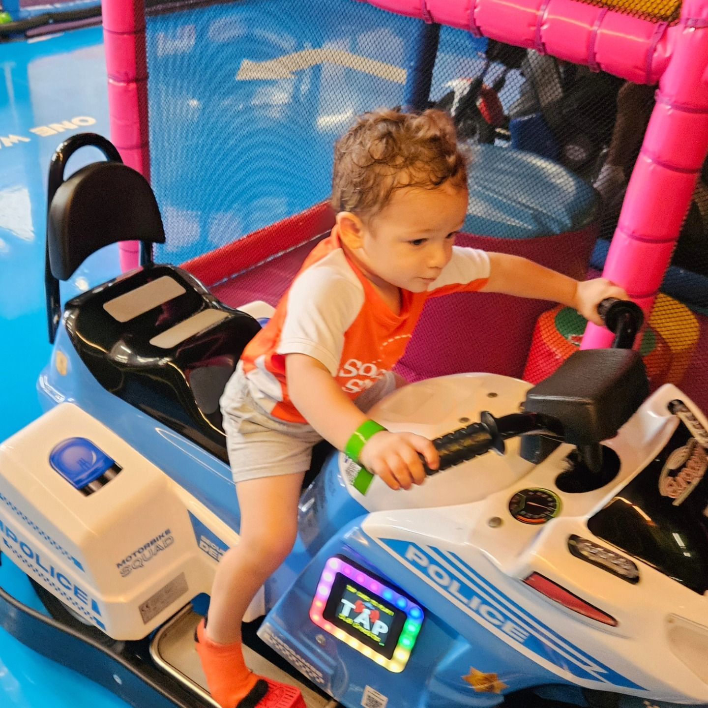 A toddler sits on a blue and white plastic police motorcycle ride inside a colorful indoor play area.