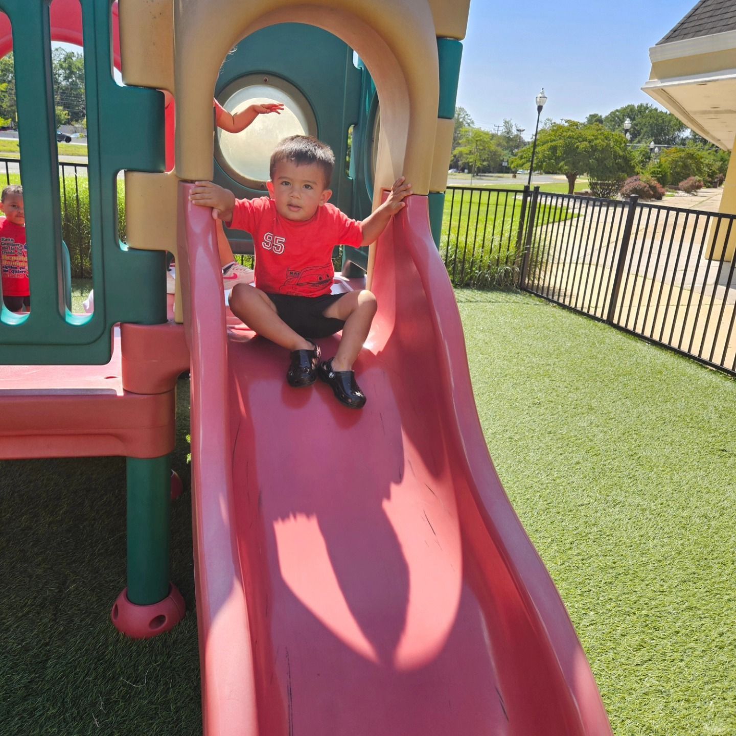A toddler in a red shirt sits at the top of a red playground slide on a sunny day.