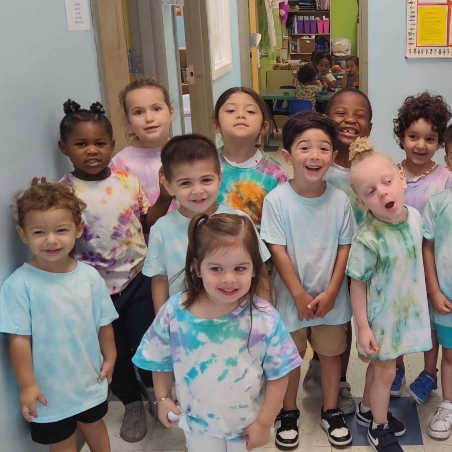 A group of children in tie-dye shirts stand together smiling indoors, likely in a classroom.