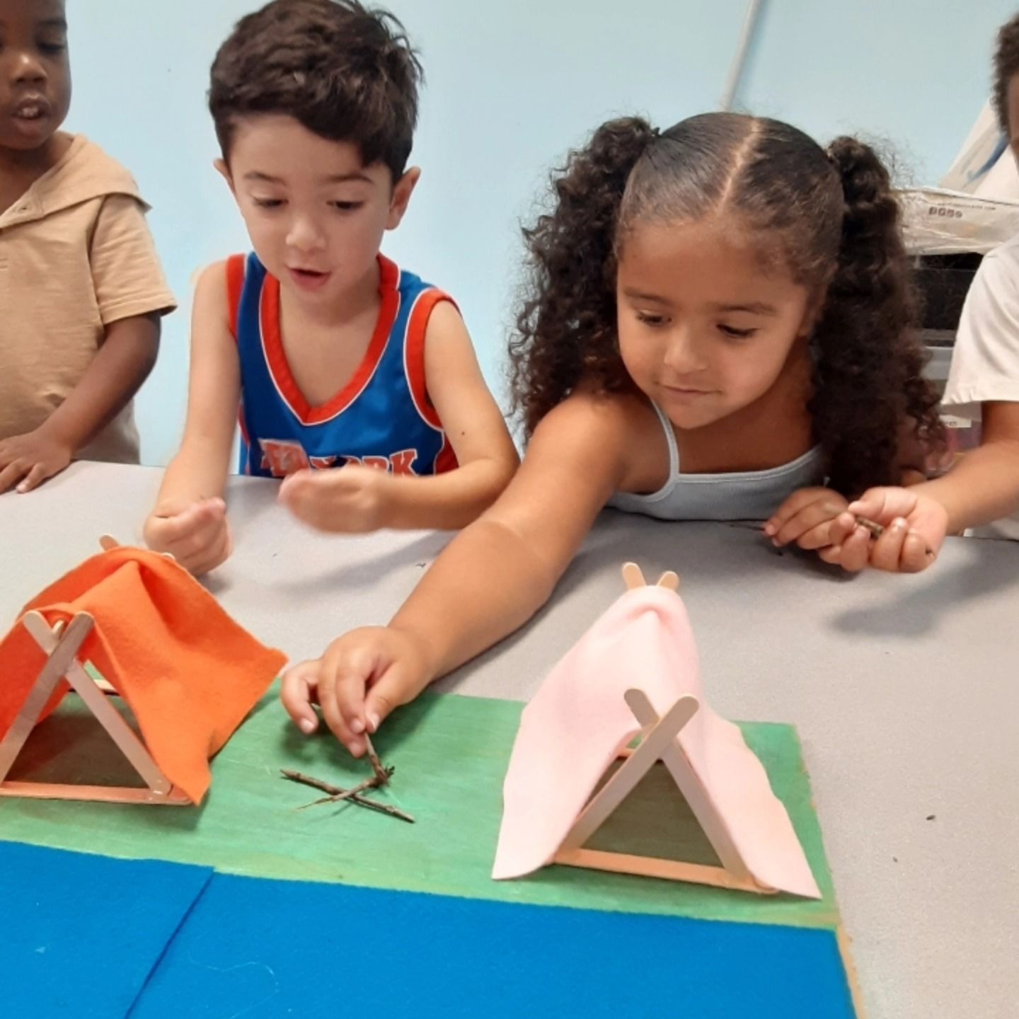 Children construct miniature tents from craft sticks and fabric on a colorful mat during an interactive activity.