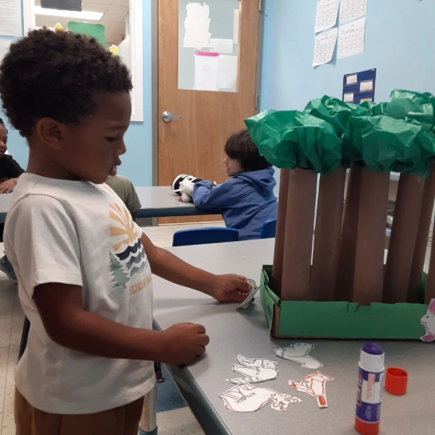 A child in a classroom crafting a forest model with cardboard tubes, green tissue paper, and animal cutouts on a table.