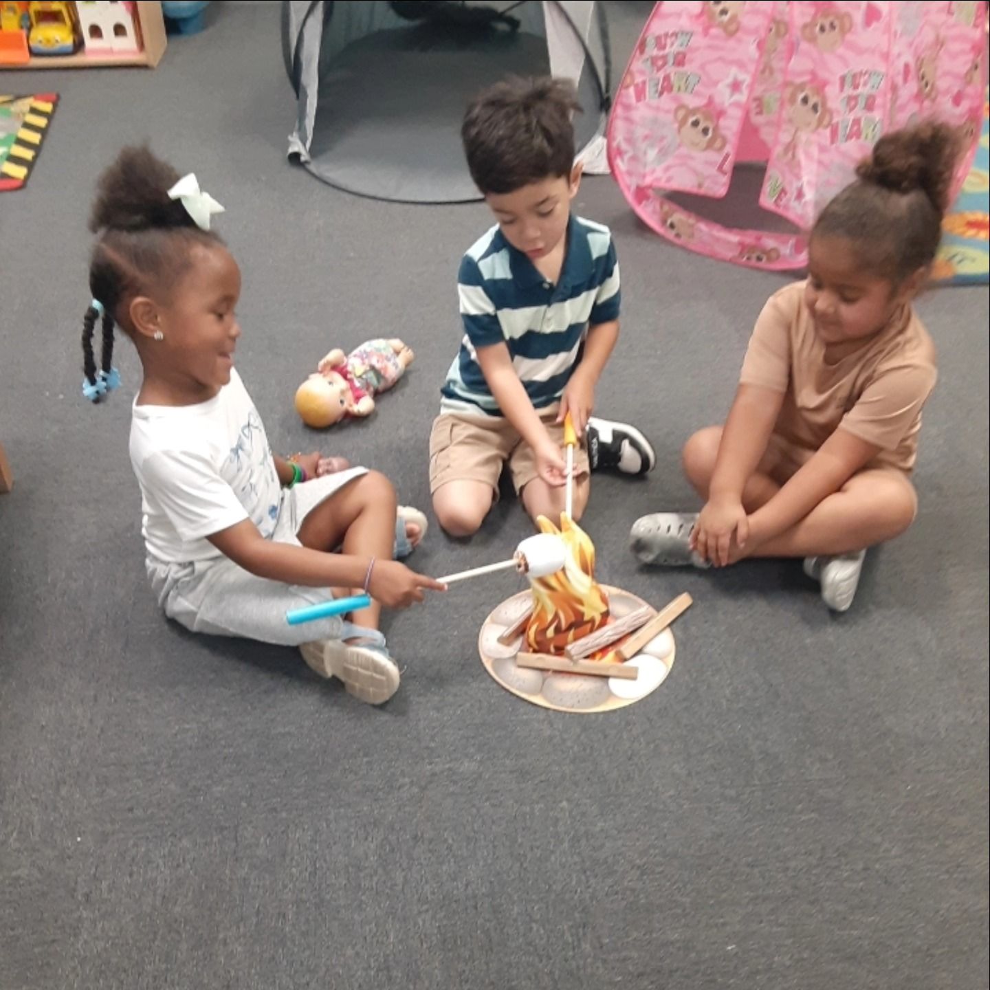 Three children sit on a rug playing with a toy campfire and roasting sticks in a classroom setting.