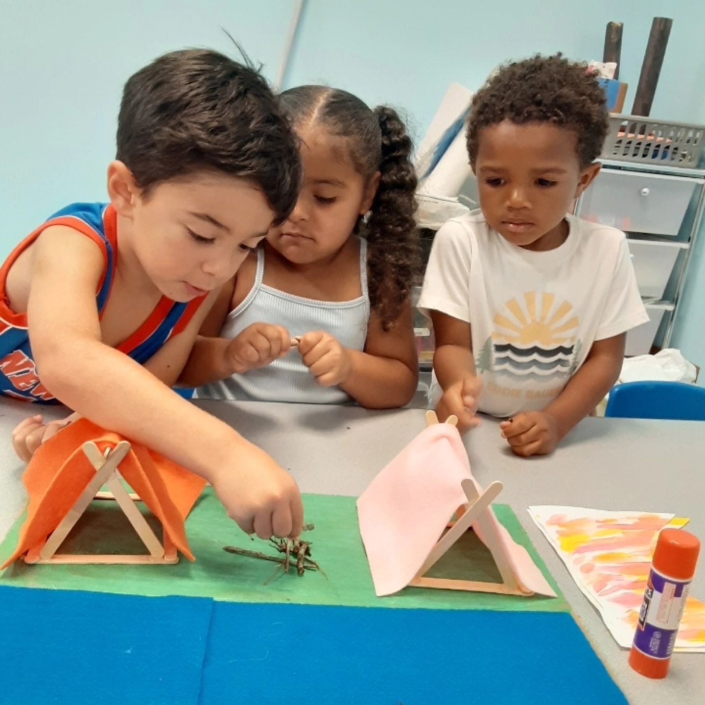 Three children build a miniature campsite on a table with popsicle stick tents, felt, and twig decorations.