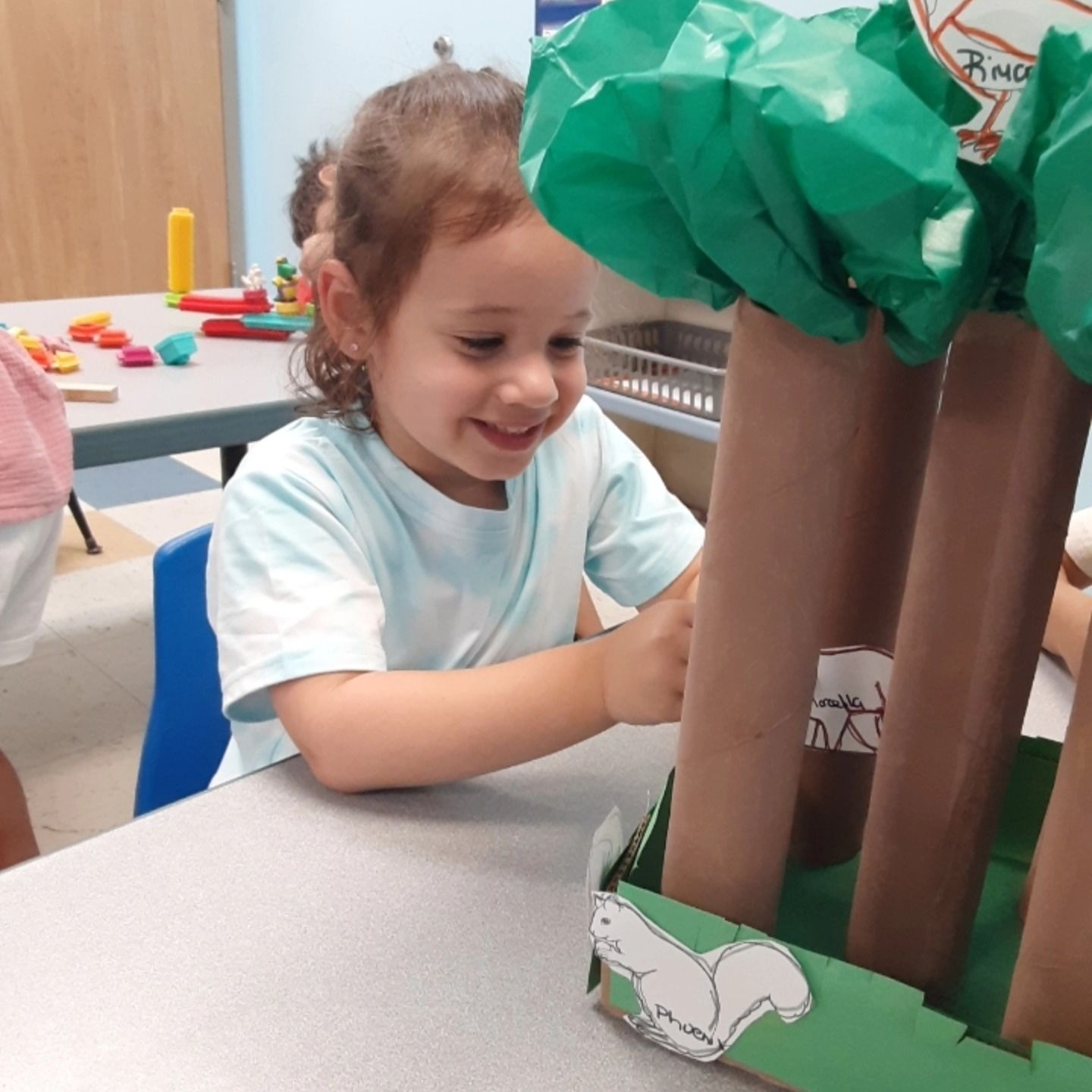 A person smiling while playing with a craft project featuring paper-towel-roll trees and animal cutouts in a classroom.