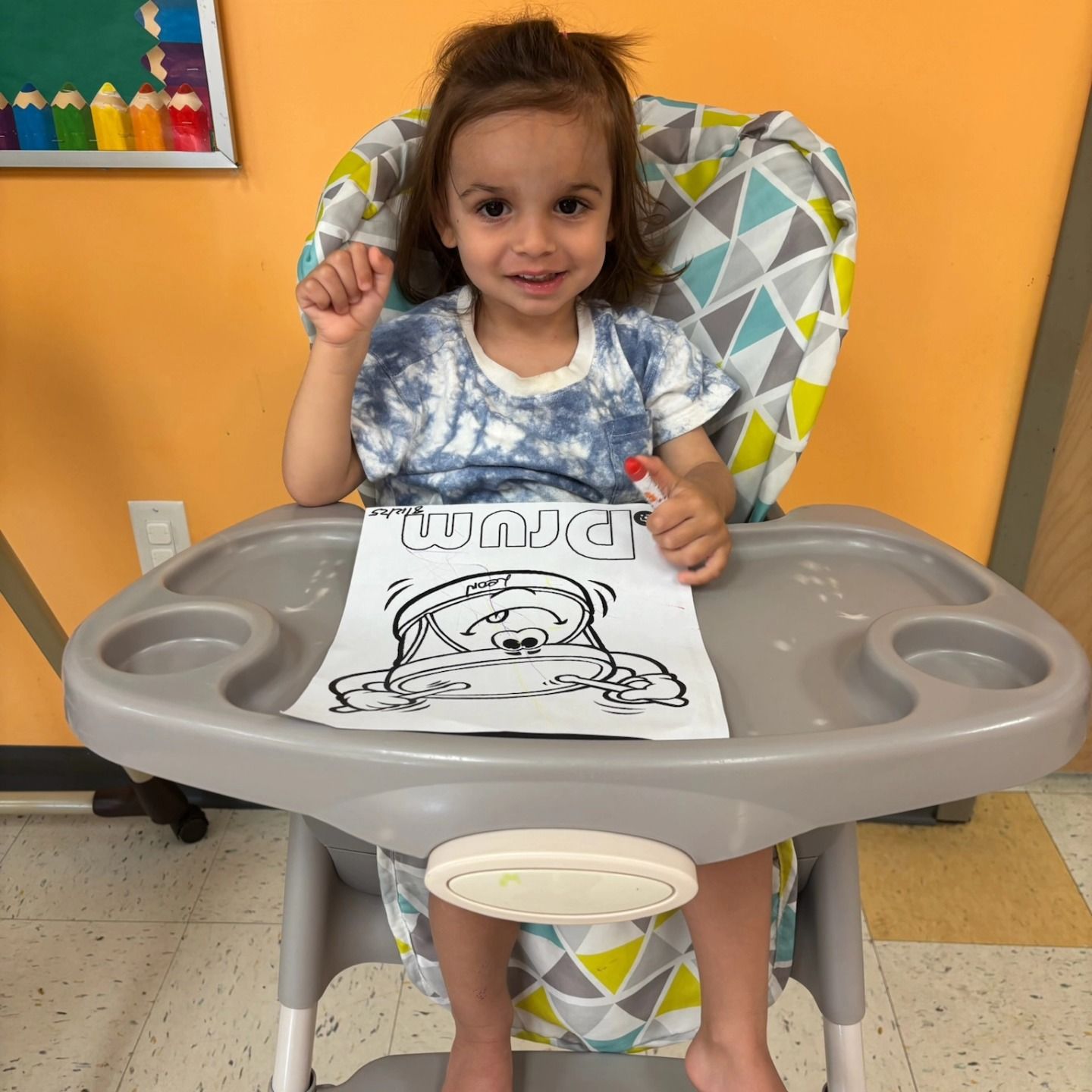 A child sits in a high chair, smiling and holding a marker over a coloring sheet with a cartoon illustration.