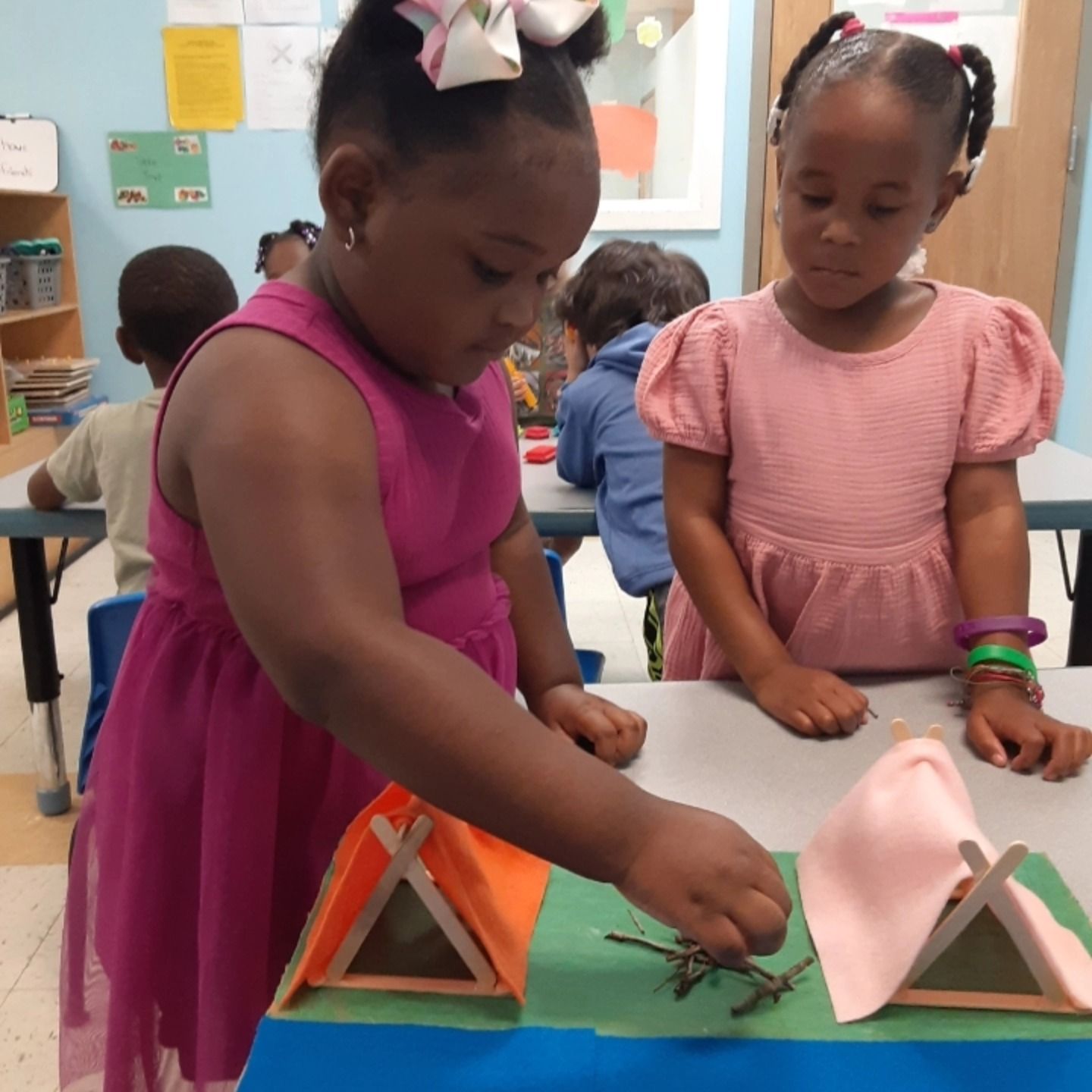 Two children in a classroom work together on a diorama with miniature tents and a pretend campfire on a table.