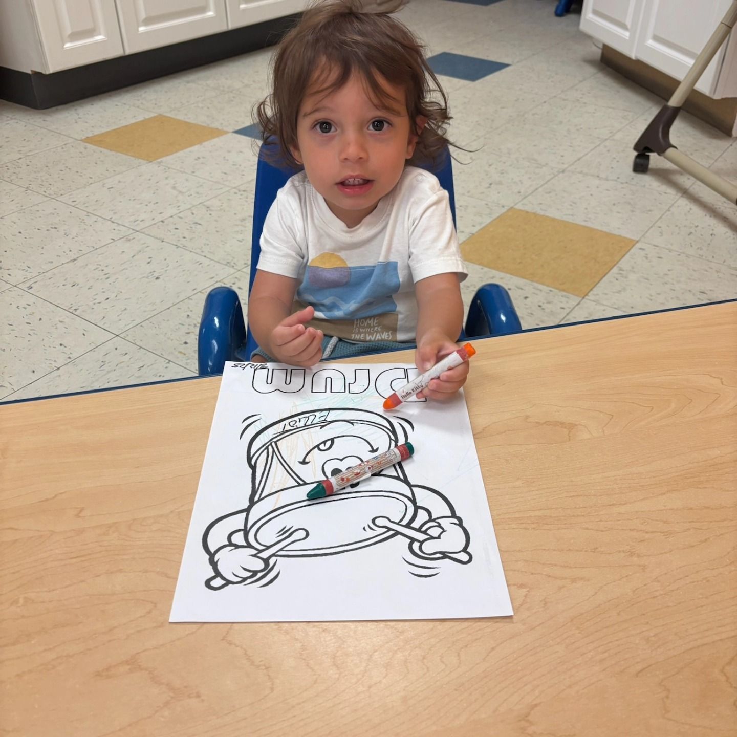 A young child sits at a desk with a drum coloring page, holding an orange crayon while a green crayon rests on the paper.