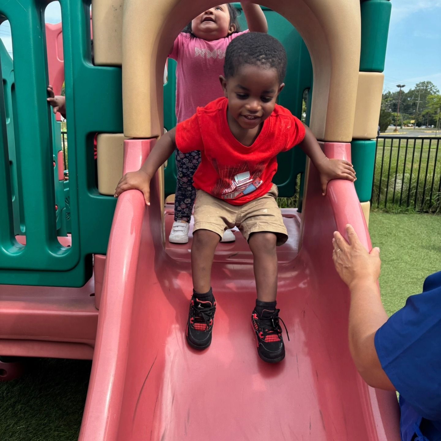 Two children slide down a pink playground slide, with an adult's hand reaching out nearby.
