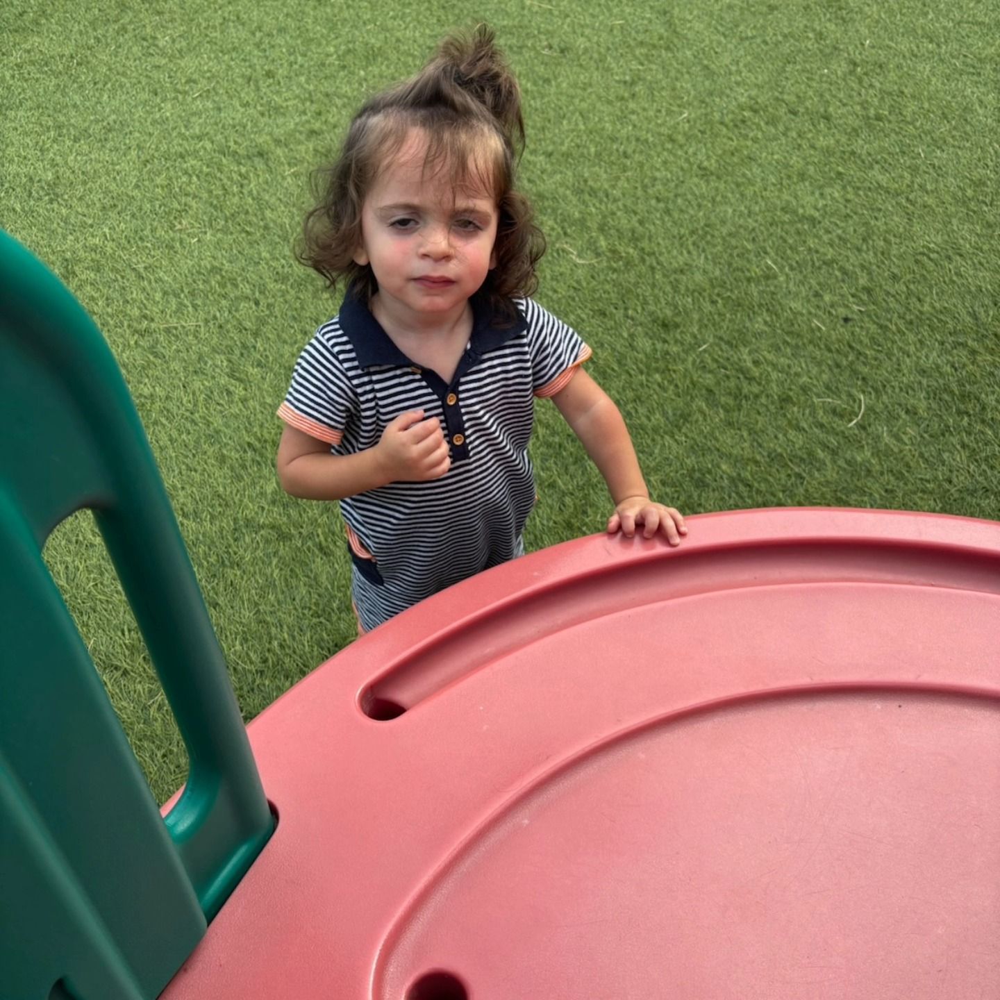 A person in a striped polo shirt stands on a grassy playground near a red plastic structure, looking toward the camera.