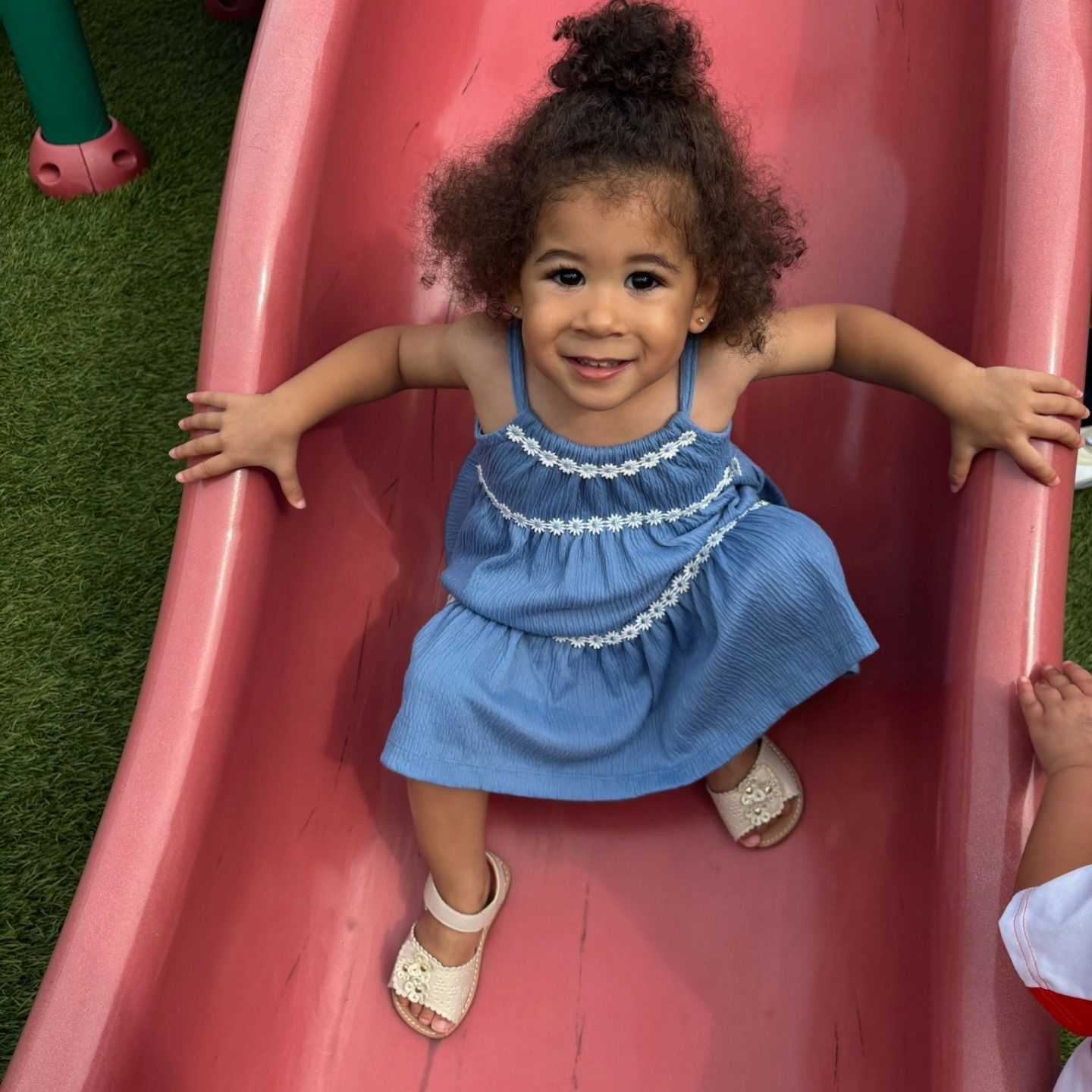 A smiling child in a blue dress with white trim sits on a pink slide outdoors.