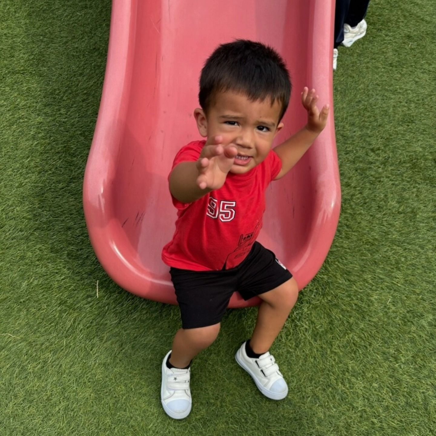 A child in a red shirt and black shorts sits on a pink slide, reaching a hand toward the camera on green turf.