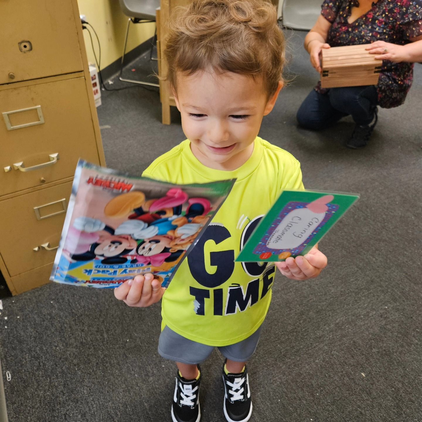 A smiling child in a bright yellow shirt holds a Mickey Mouse activity pack and a small card in an office setting.