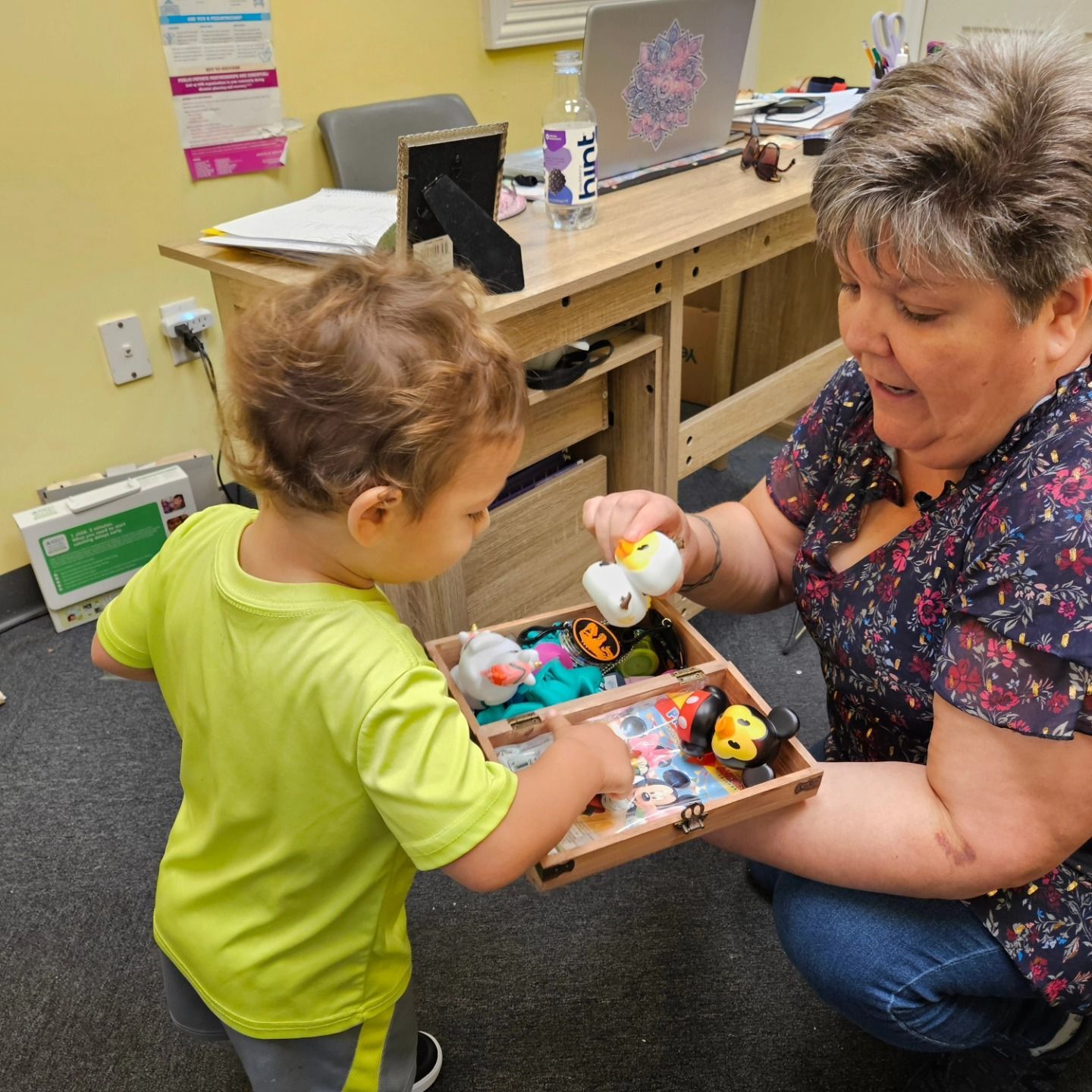 An adult and child interact while holding a small wooden box filled with colorful small toys in an office setting.