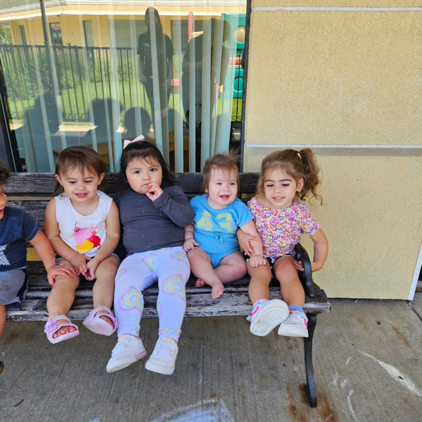 Five children sit side-by-side on a rustic outdoor bench against a pale wall, smiling and posing for a group photo.