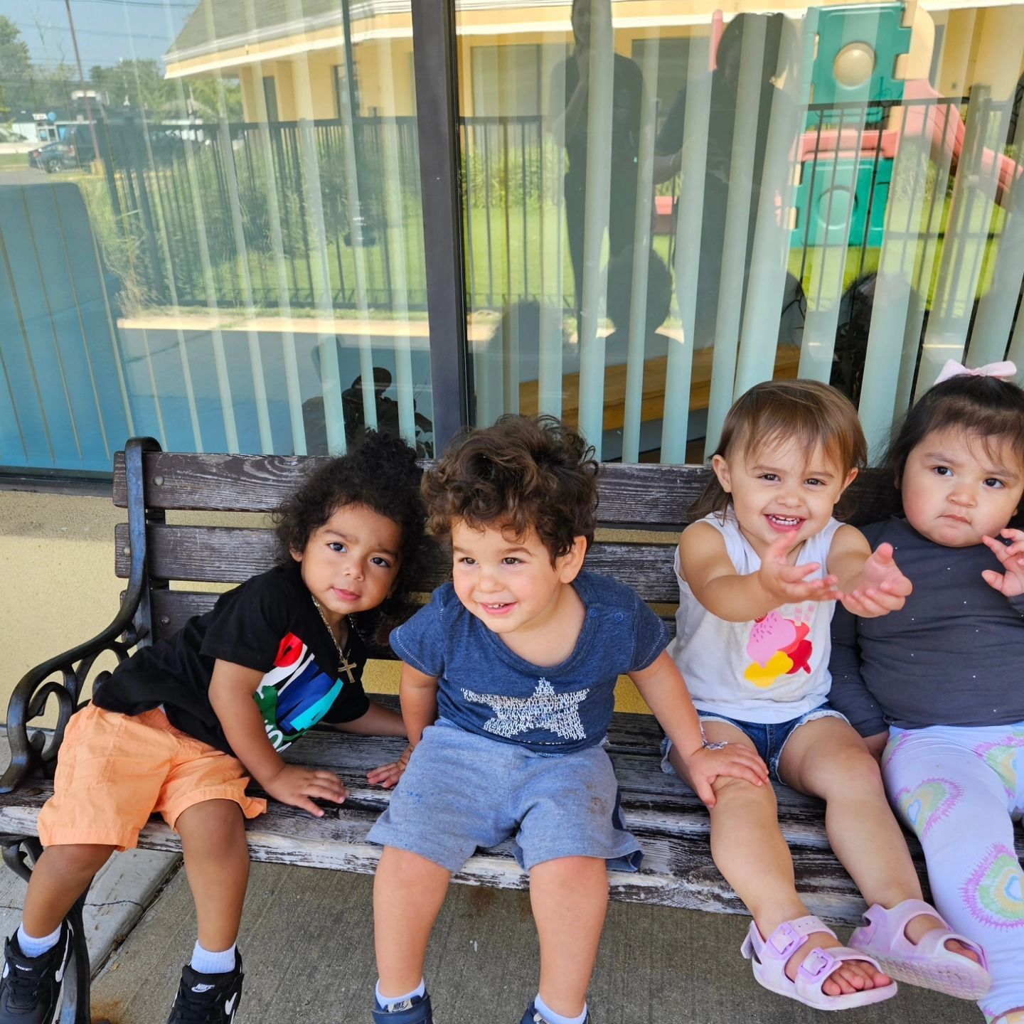 Four children sit side-by-side on a wooden bench outdoors, smiling and reaching toward the camera.