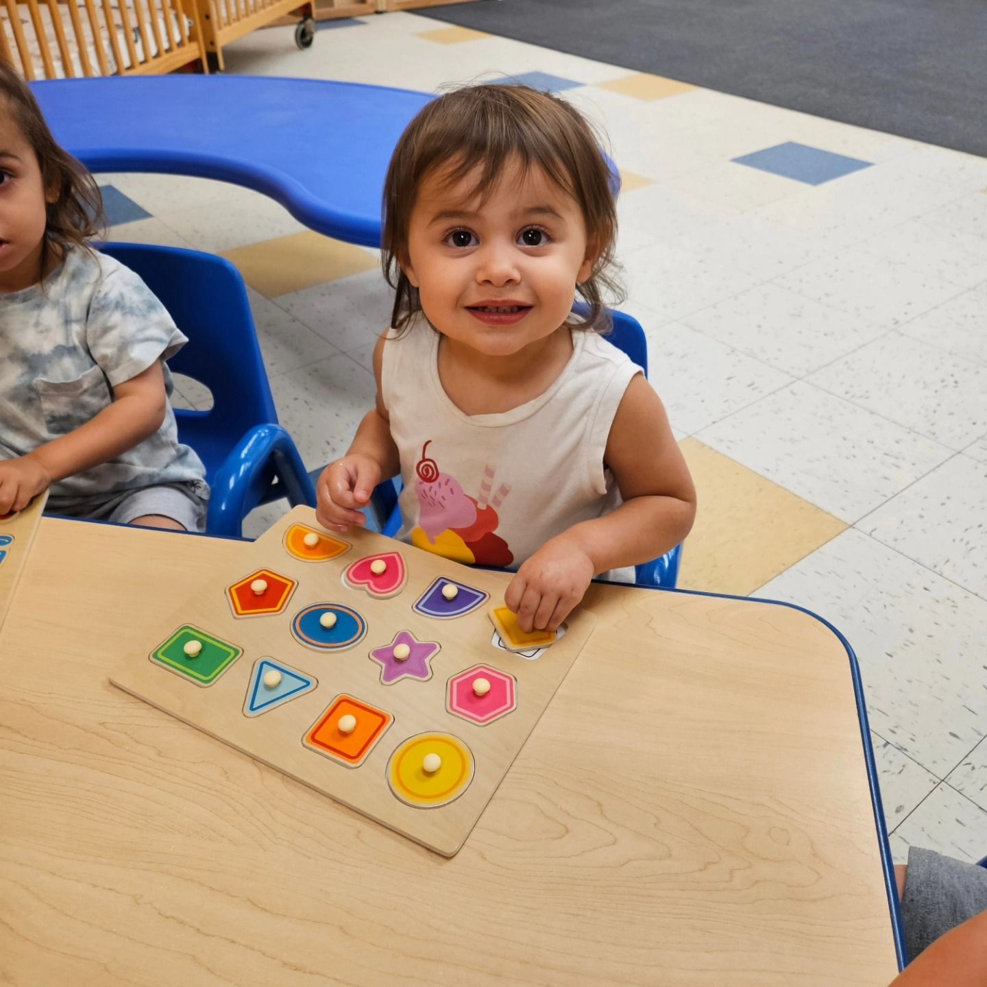 A child sits at a table playing with a colorful wooden shape-sorting puzzle in a classroom setting.