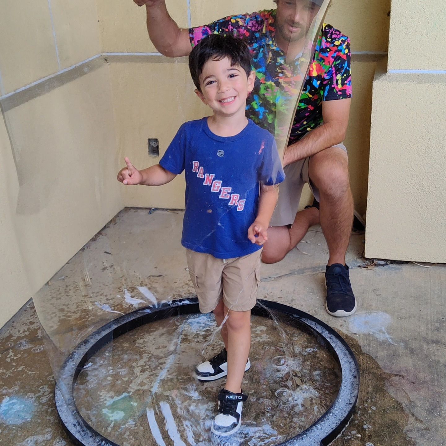 A smiling child stands inside a large, circular bubble wand frame as an adult lifts it, surrounded by soap bubbles.