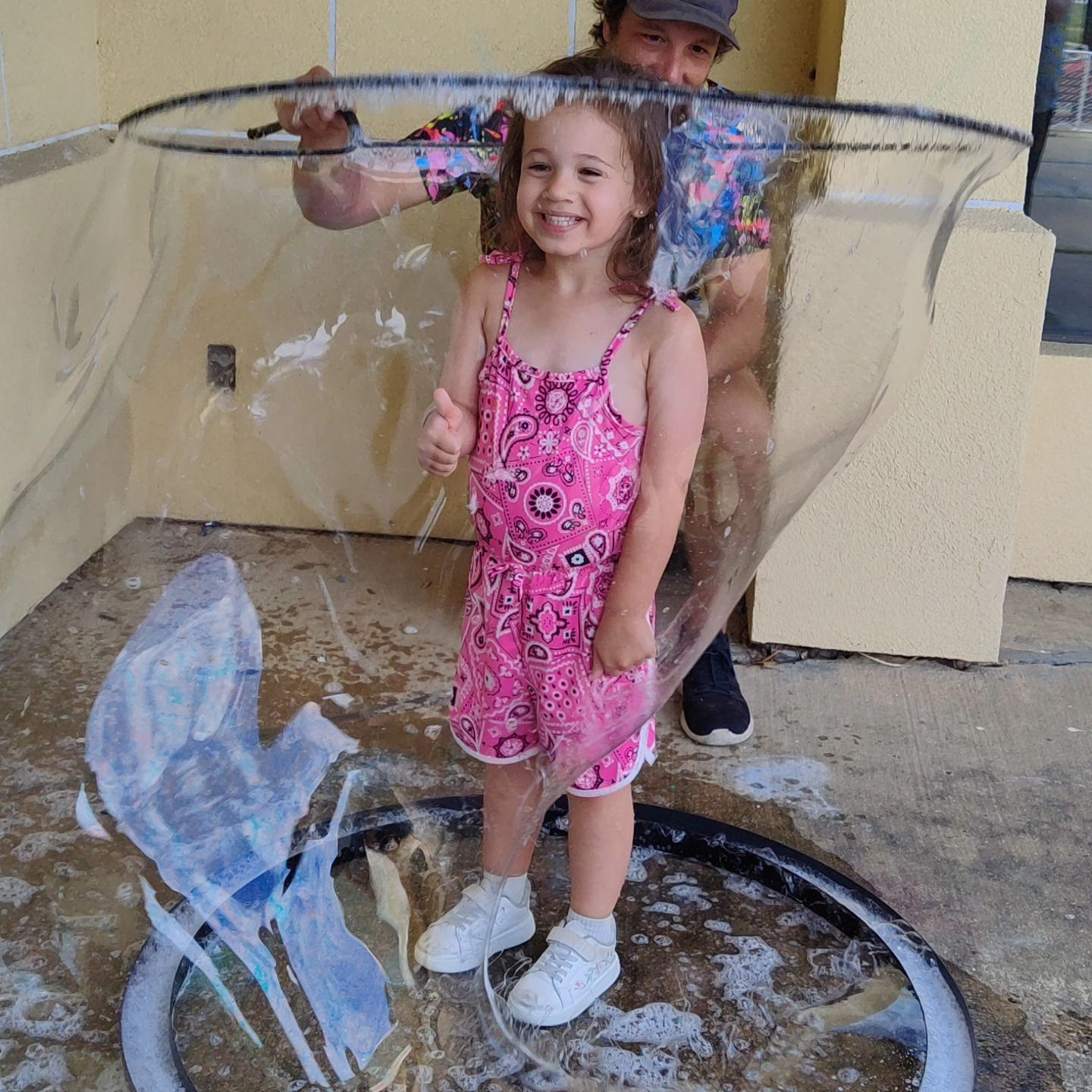 A child in a pink patterned romper stands inside a large, clear bubble held by an adult in a patterned shirt.