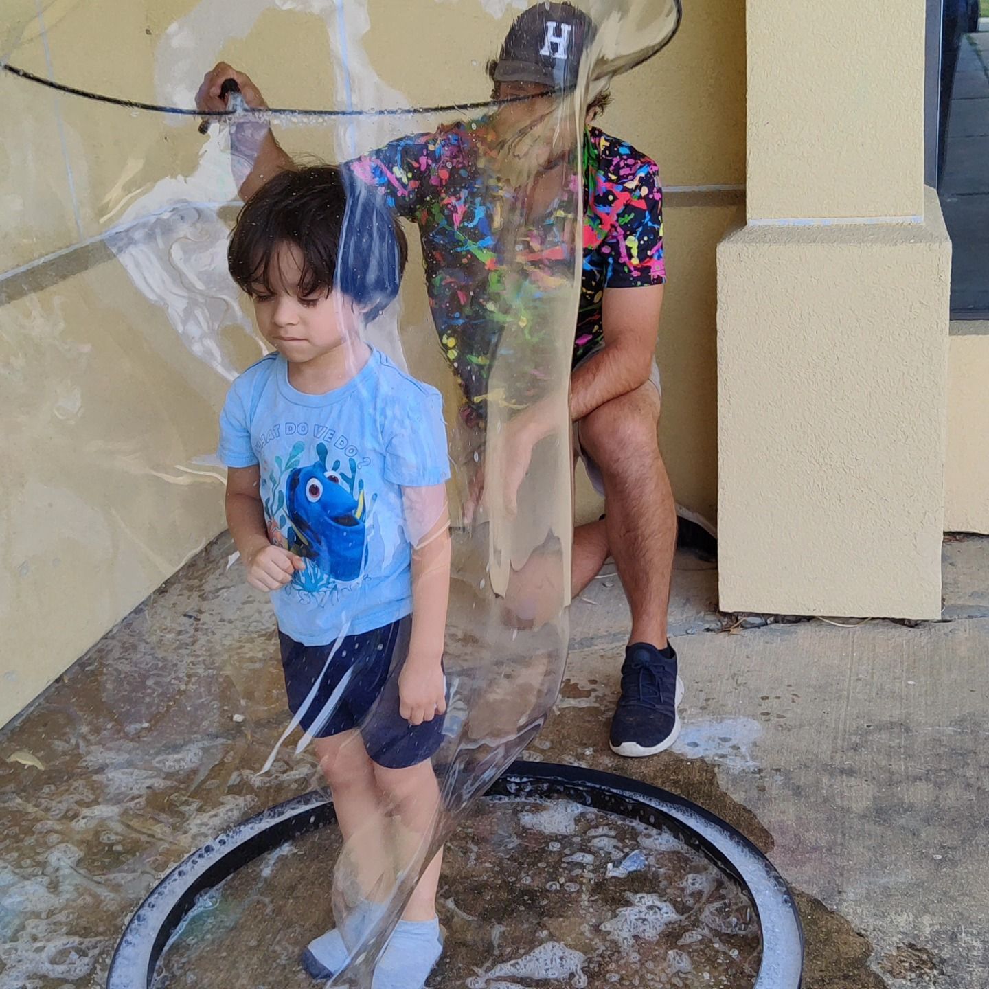 A person stands behind a child, pulling a large bubble wand upward to encase the child inside a tall, clear soap bubble.