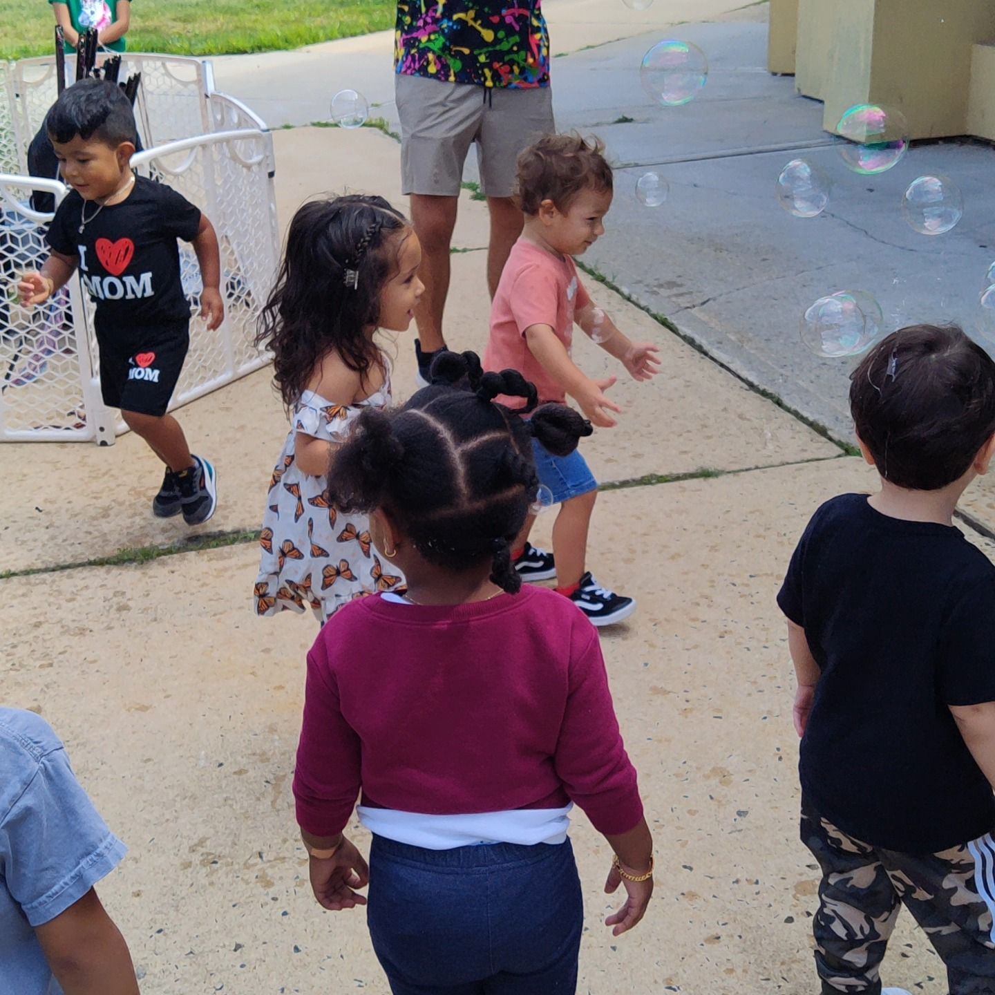 Children play outdoors with bubbles on a paved surface, some moving towards the camera while others face away.