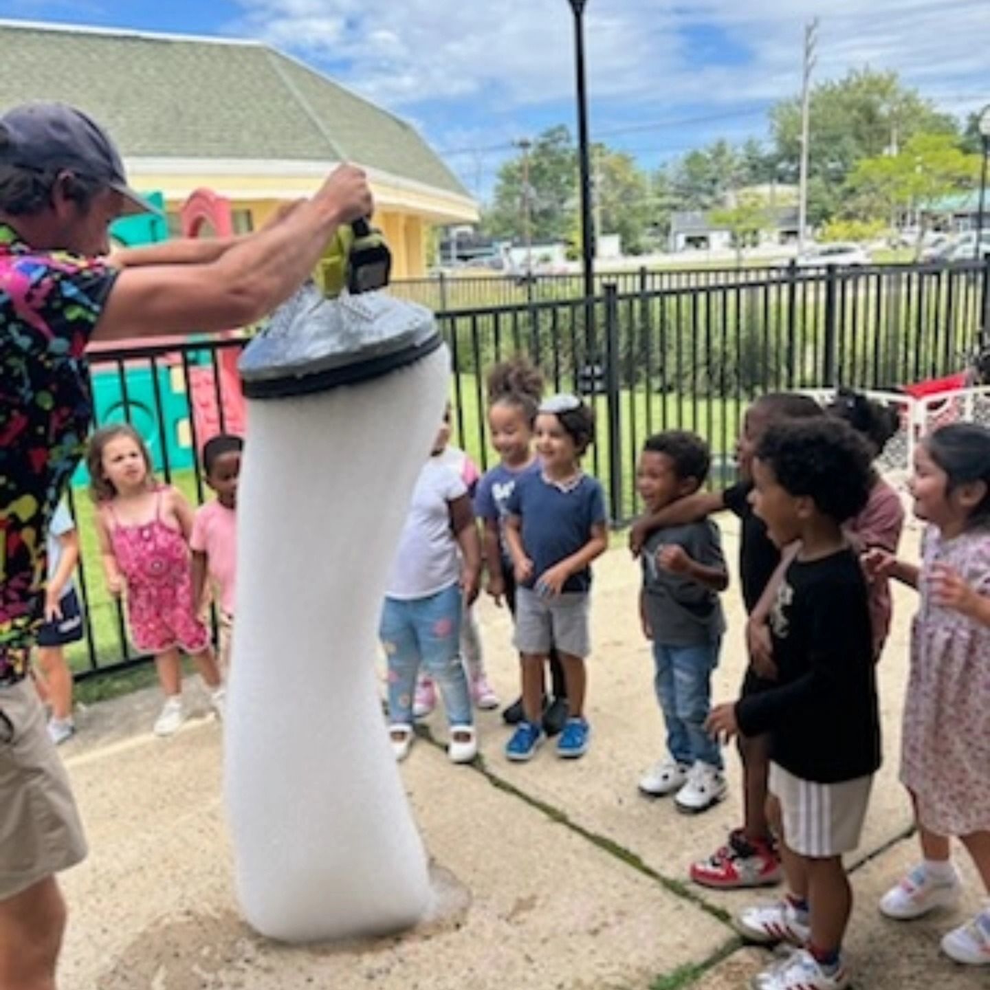 An adult uses a foam-producing tool to create a tall, white bubble tower for a group of children outdoors.