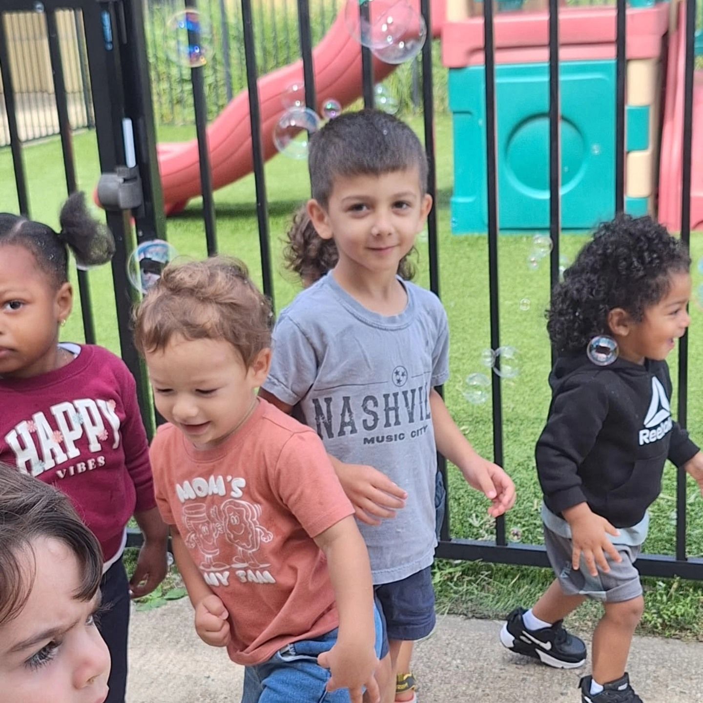 A group of children plays outdoors on a playground near a black fence as bubbles float around them.
